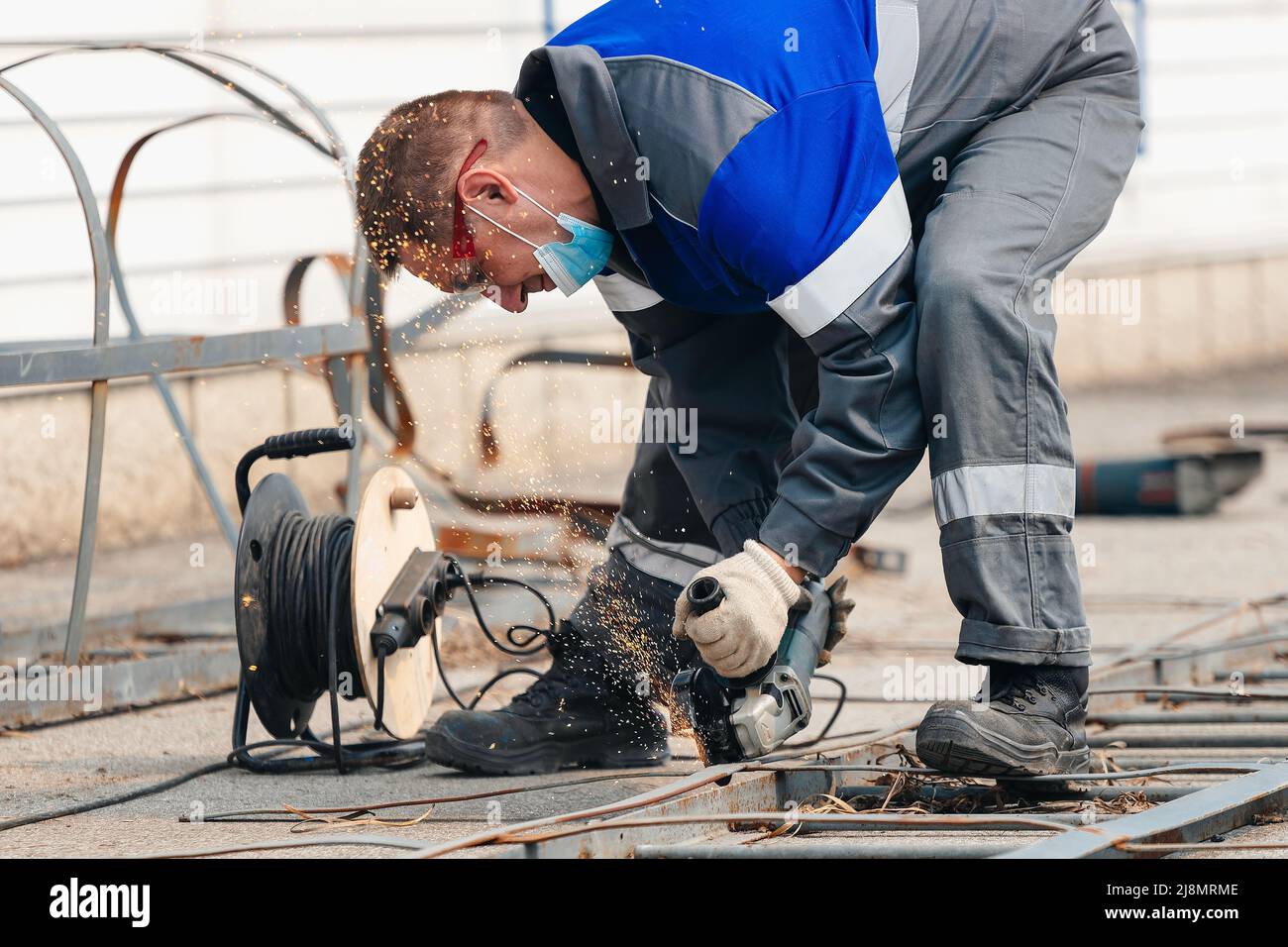 Builder in Overalls lehnt sich über und schneidet Metallblech mit Winkelschleifer und Funken fliegen. Arbeiter mit Brille bei der Arbeit auf der Straße. Authentischer Workflow. Stockfoto