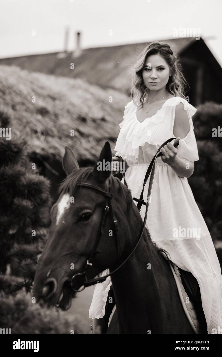 Eine Frau in einer weißen Sundress auf einem Pferd in der Nähe einer Farm. Schwarz-Weiß-Foto Stockfoto