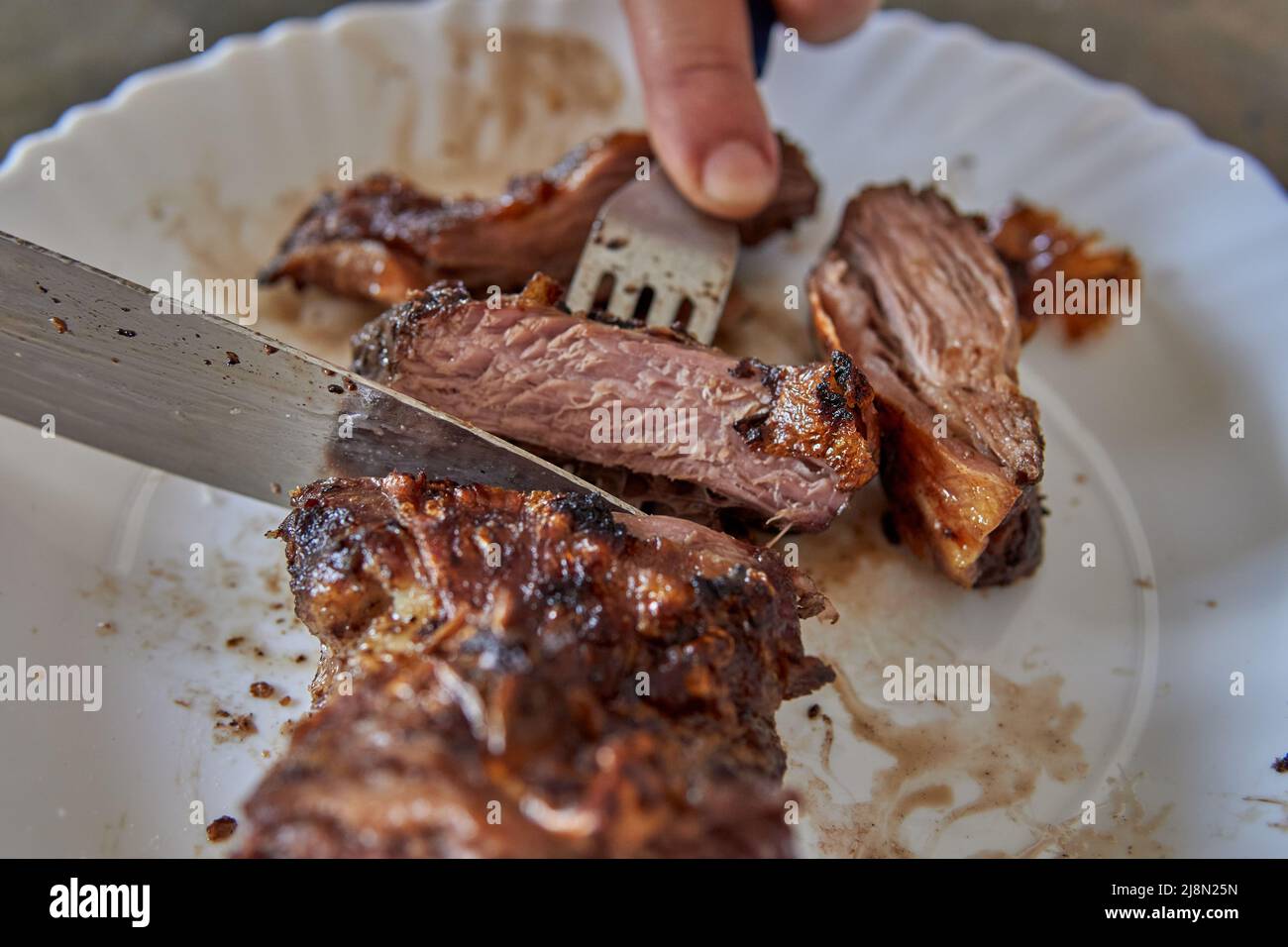 Argentinischer Grill. Menschenhände schneiden ein Stück Fleisch mit Messer und Gabel. Nahaufnahme von gegrilltem Rindersteak in Scheiben auf einem Teller Stockfoto