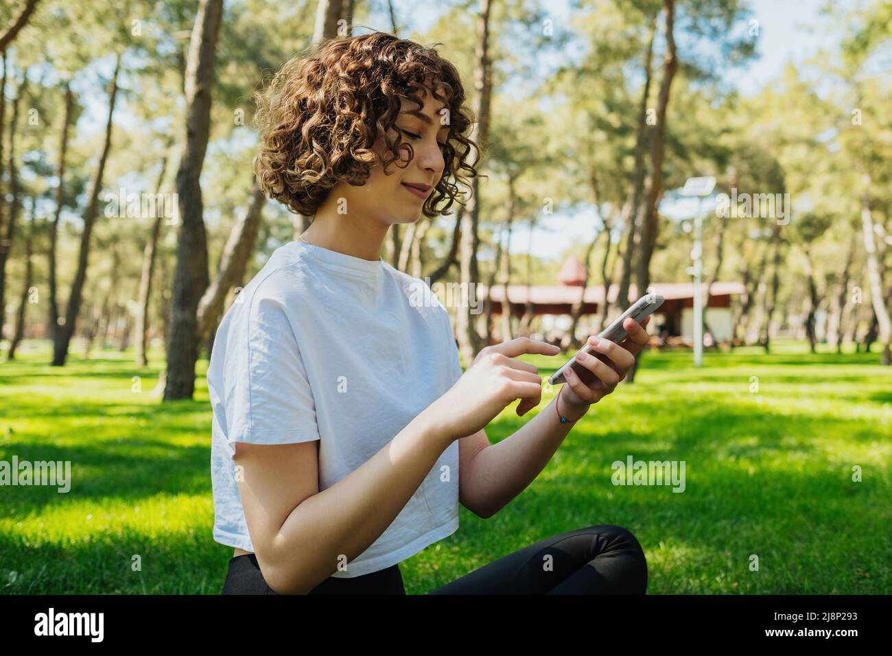 Halblange Körpergröße Foto rothaarige Frau trägt weißes T-Shirt auf Gras sitzen lächelnd glücklich mit Smartphone Surfen im Internet oder Messaging mit Freund Stockfoto