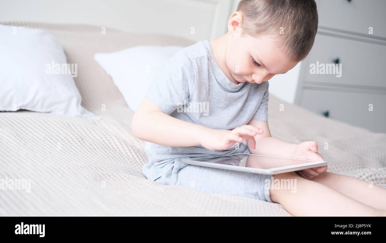 Ein kleiner kaukasischer Junge in einem hellen Raum in einem grauen T-Shirt spielt mit einem Tablet, während er auf der Couch sitzt. Stockfoto