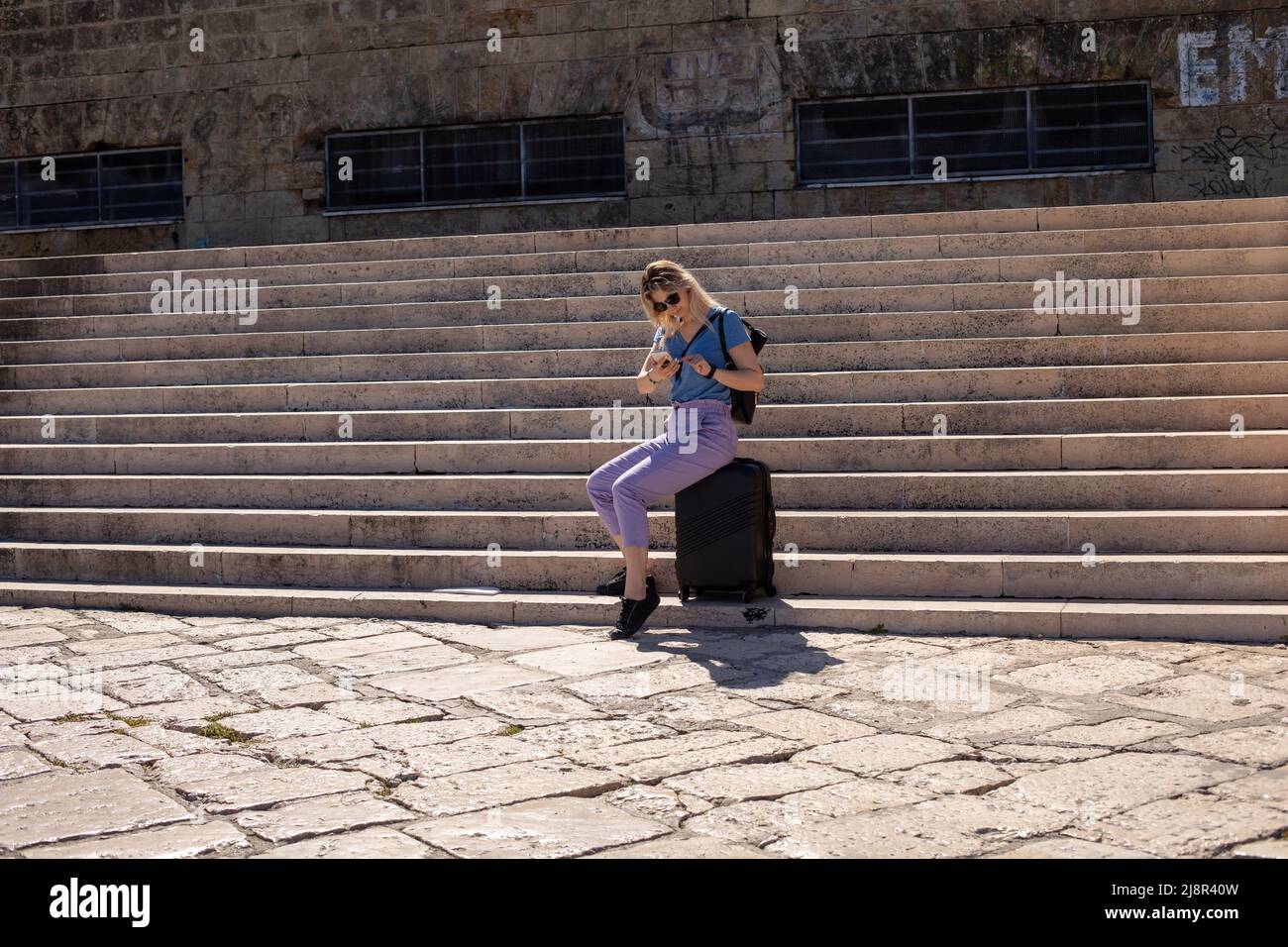 Eine blonde Frau mit Sonnenbrille sitzt auf dem Gepäck auf einer großen Treppe in der Nähe eines Steingebäudes. Suche nach elektronischen Tickets und Karten Stockfoto