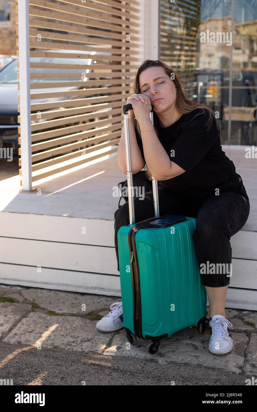 Vertikale, schwülige blonde Frau in legerer Kleidung mit Gepäck, auf Taschen gelehnt. Sit Bahnhof, wartet auf Transport Stockfoto