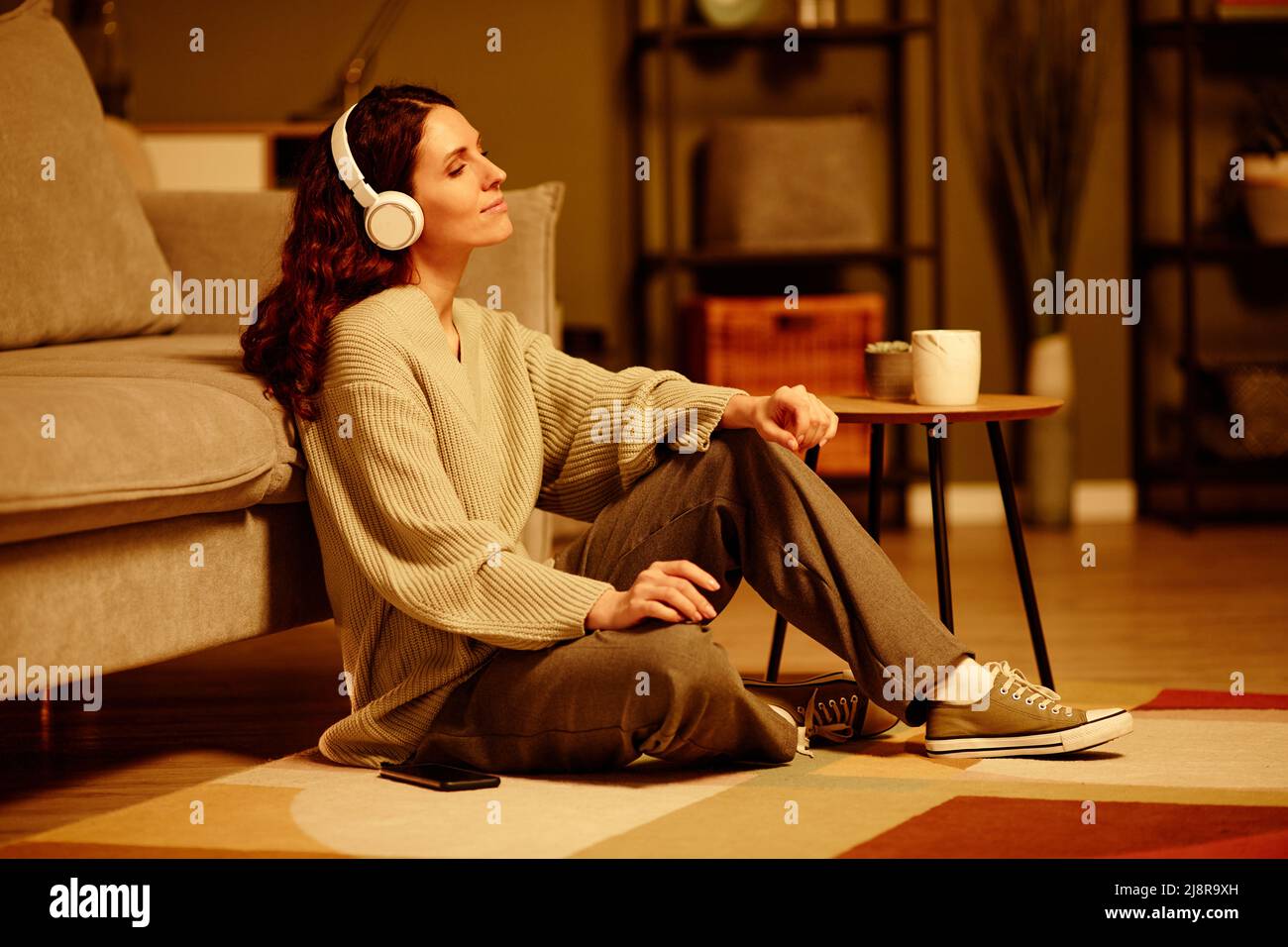 Elegante kaukasische Frau in legerer Kleidung, die abends entspannt auf dem Boden im Wohnzimmer sitzt und Musik über kabellose Kopfhörer hört Stockfoto