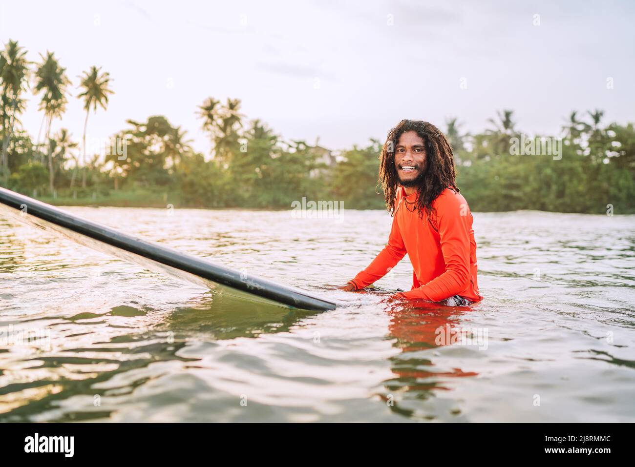 Schwarzer langhaariger Teenager-Mann, der auf einem langen Surfbrett schwimmt und auf eine Welle wartet, die zum Surfen bereit ist, mit Palmenhain-Sonnenstrahlen. Extreme Wassersportarten o Stockfoto