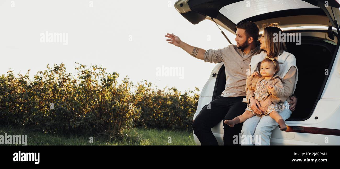 Junge kaukasische Familie sitzt im Kofferraum des SUV-Autos, Mama und Papa mit Baby-Tochter Spaß am Wochenende im Freien, Copy Space Stockfoto