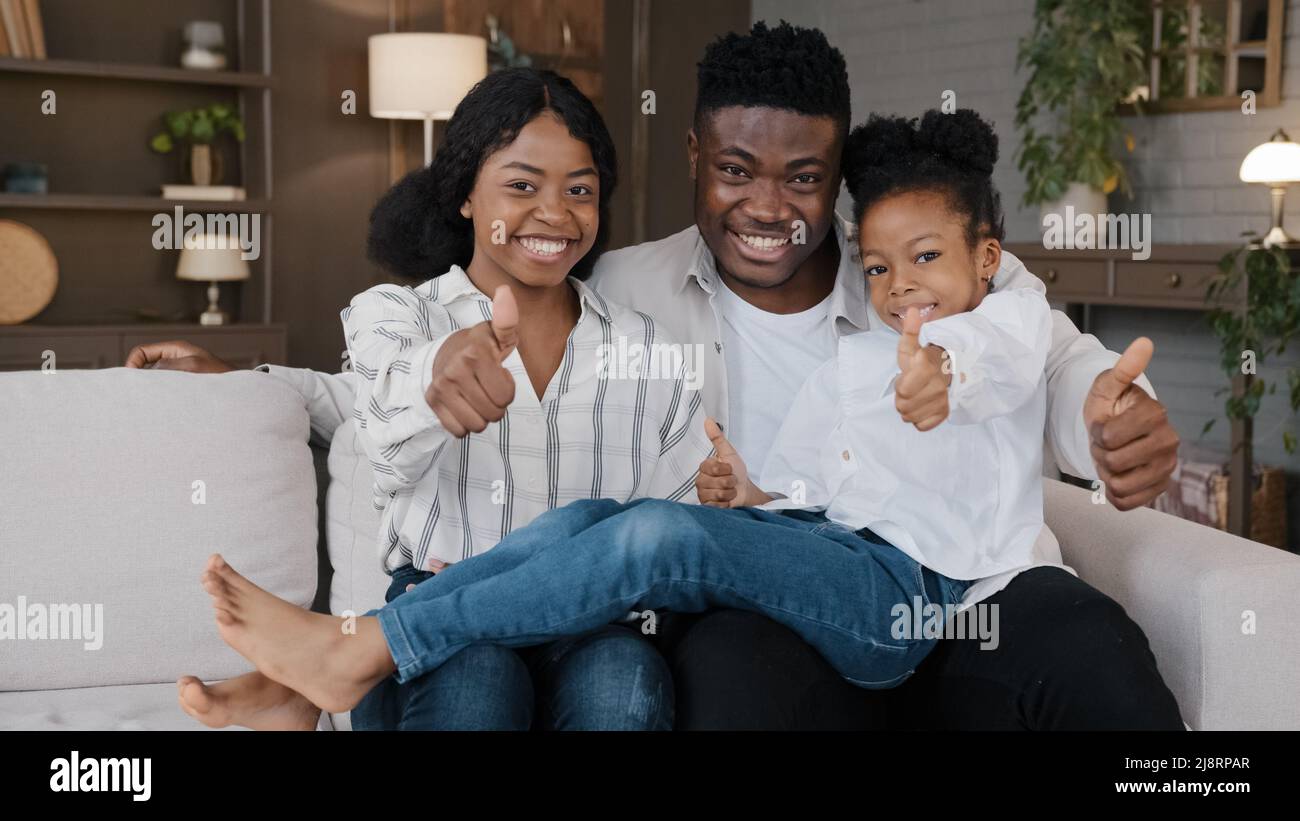 Glückliche afrikanische Familie junge Vormund Eltern und kleine Tochter adoptierte Mädchen Kind sitzt auf dem Sofa zu Hause eigene Wohnung neues Haus Blick auf Kamera s Stockfoto