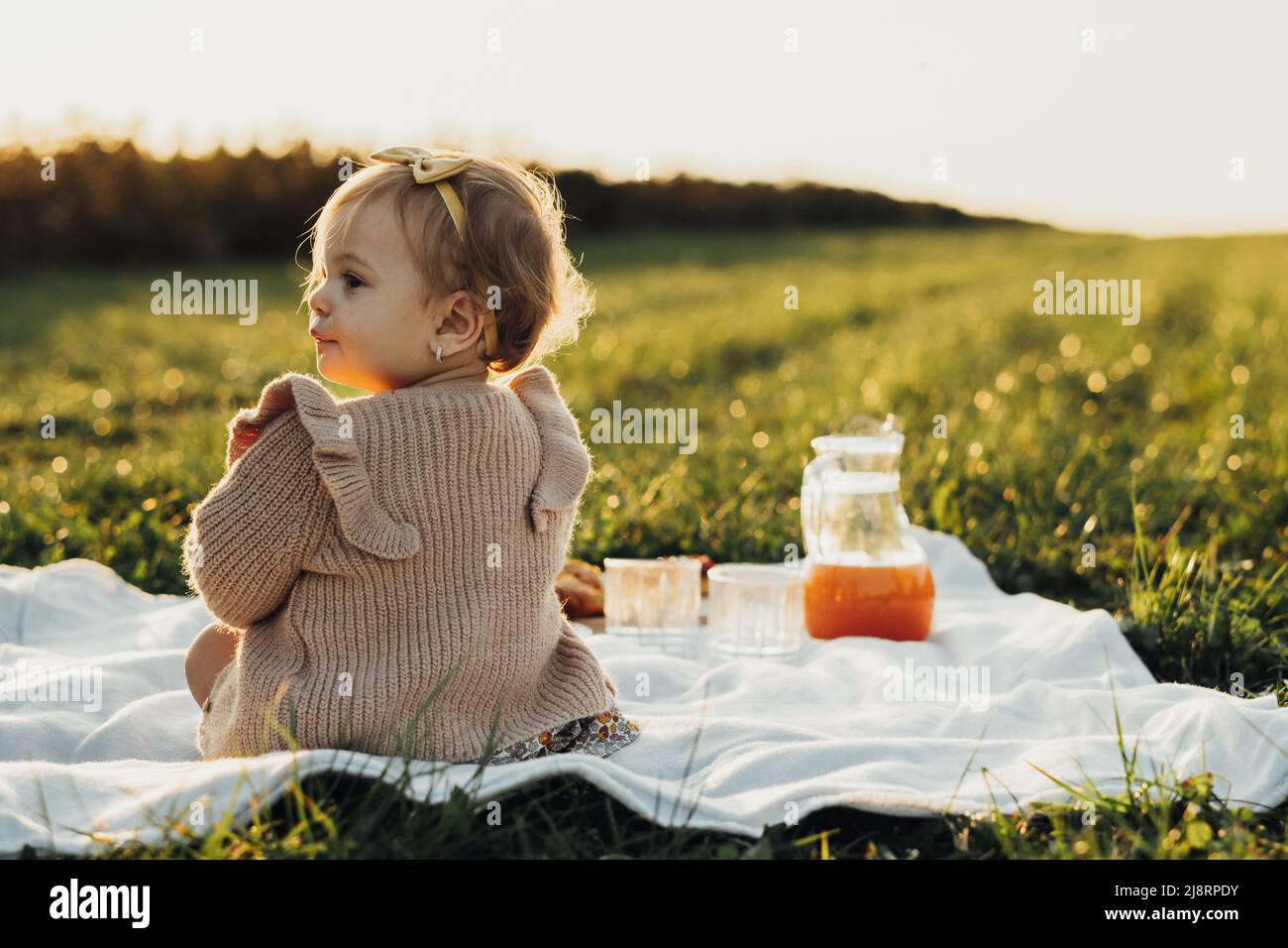 Rückansicht des kleinen Mädchens, das auf einem Plaid im Freien bei Sonnenuntergang sitzt Stockfoto