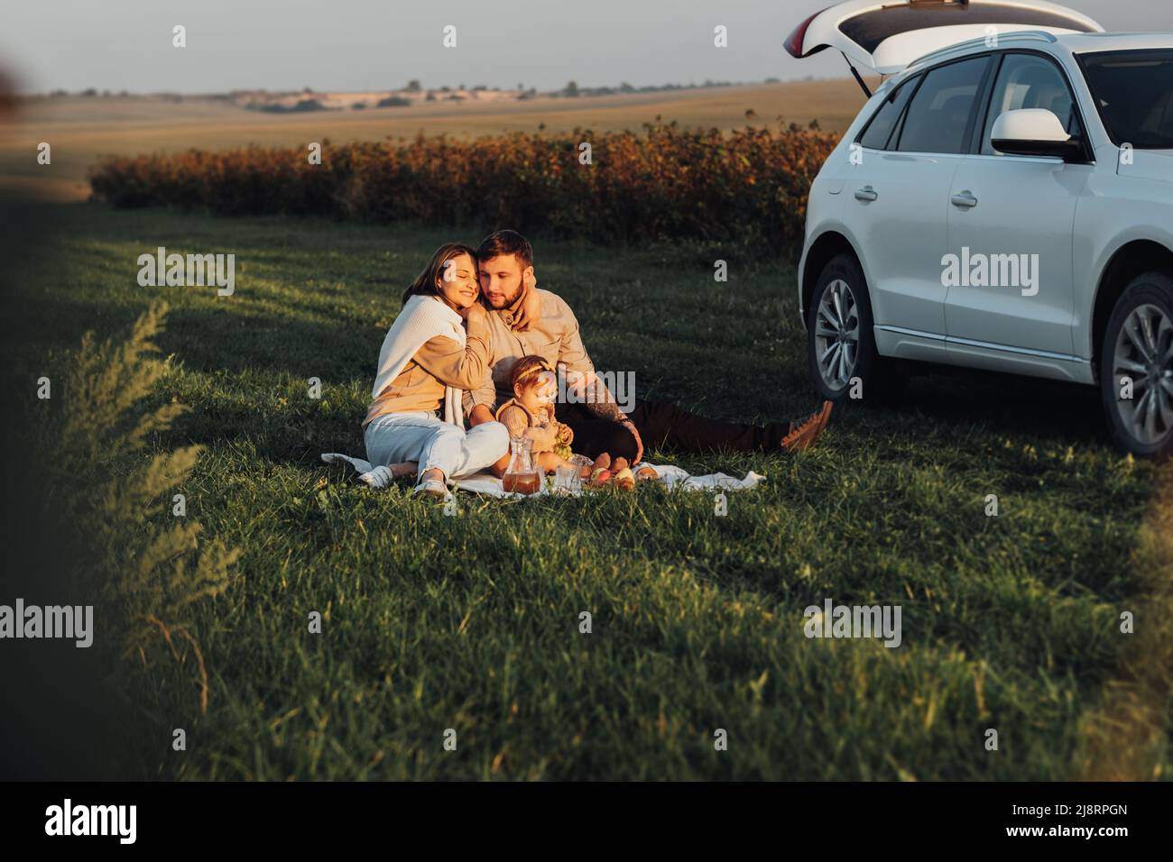 Fröhliche junge Familie, die mit einem Geländewagen unterwegs ist, und Mama Papa und ihre kleine Tochter, die am Wochenende im Freien bei Sonnenuntergang ein Picknick machen Stockfoto