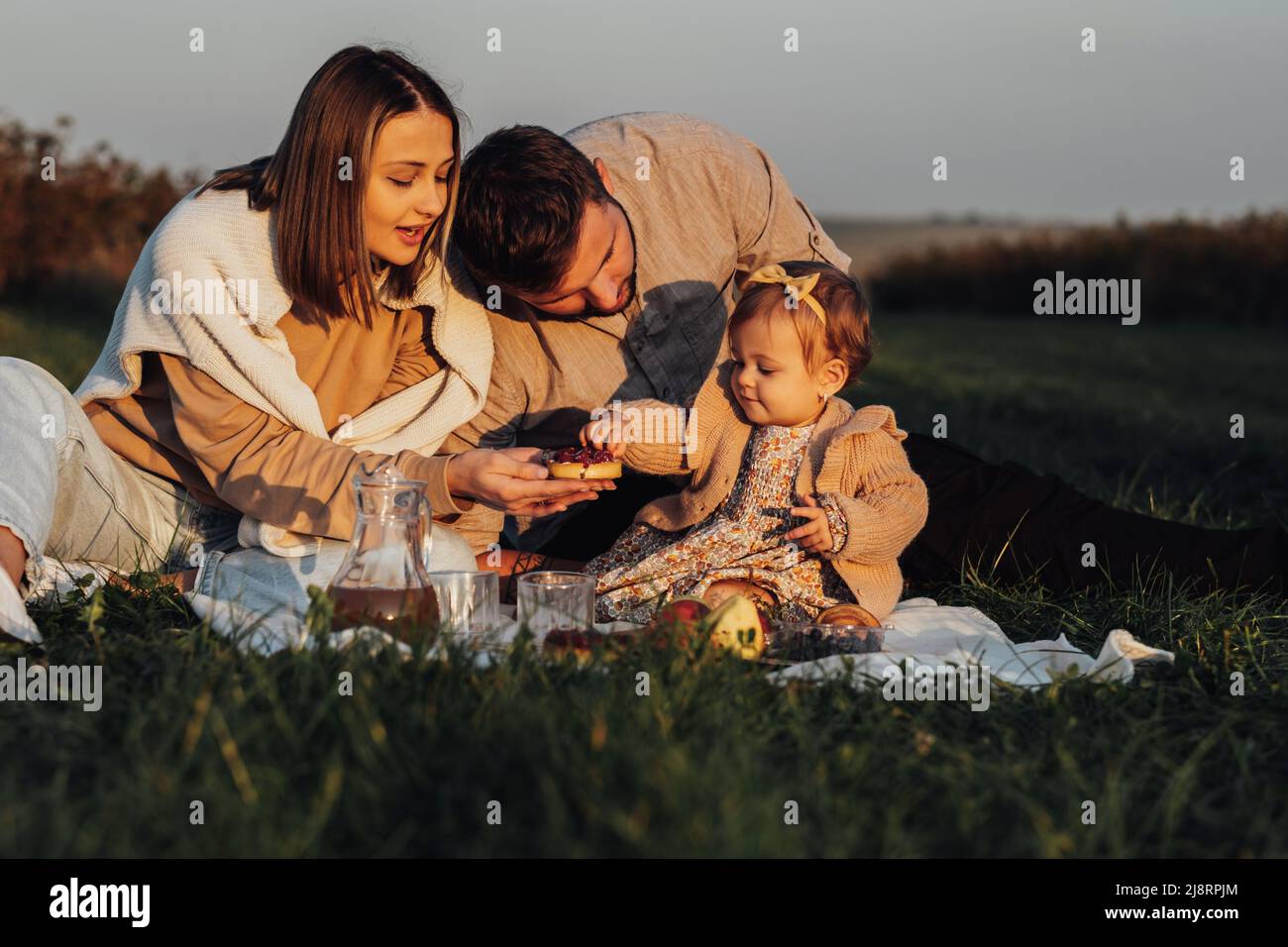 Junge Familie genießt Picknick im Freien bei Sonnenuntergang, Mama und Papa mit Baby-Tochter am Wochenende Stockfoto