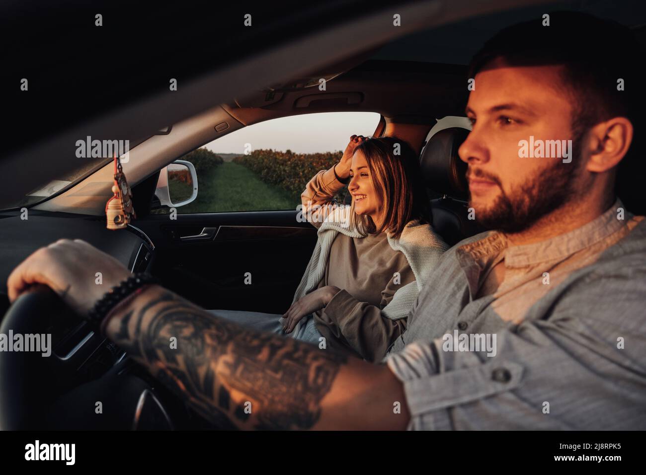 Tätowierte kaukasische Mann und junge Frau sitzen im Auto bei Sonnenuntergang, Paar genießen Road Trip am Wochenende Stockfoto