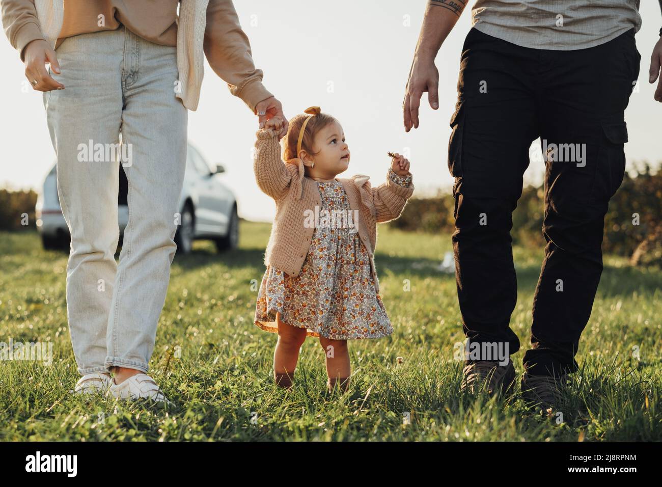 Kleine Tochter mit Eltern, die bei Sonnenuntergang im Freien auf dem Feld spazieren gehen, kleines Mädchen mit Mutter und Vater, die Zeit mit der Familie haben Stockfoto