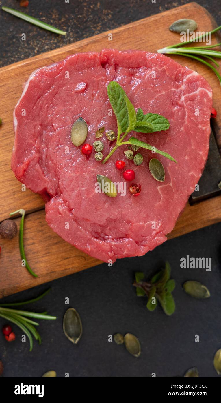 Rohes Rinderfiletsteak auf Holzbrett auf dunkler Tischansicht. Portioniertes Filet Mignon mit Gewürzen und Kräutern. Konzept der Sommerkost Stockfoto
