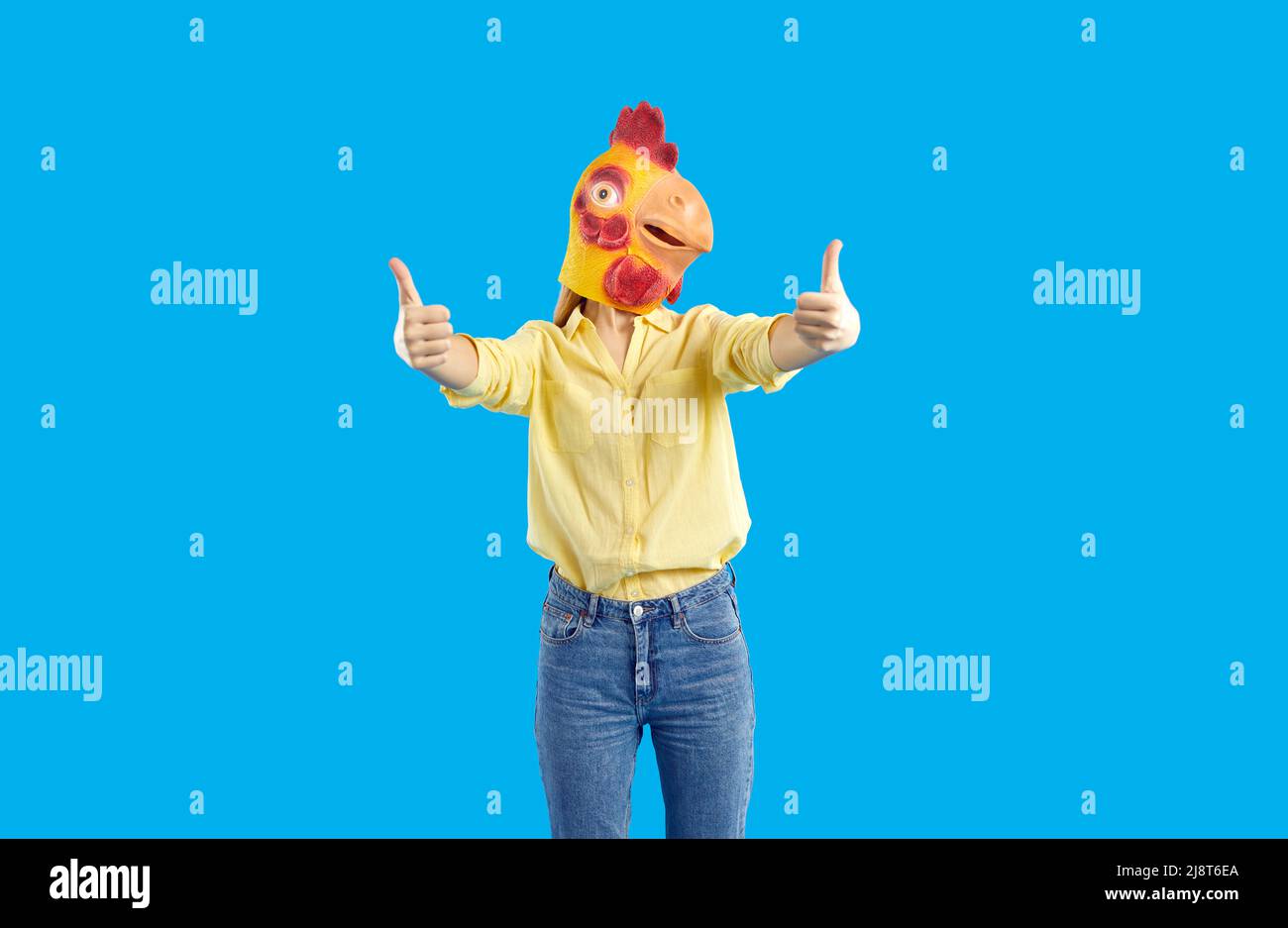 Fröhliche lustige und humorvolle Frau in Gummimaske aus Huhn zeigt Daumen nach oben auf blauem Hintergrund. Stockfoto