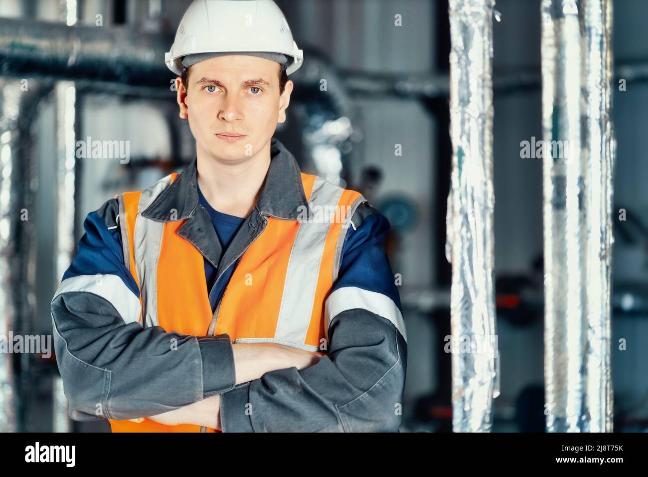 Porträt eines jungen Schwerindustrieingenieurs in Schutzweste und Arbeitskleidung und Helm. Selbstbewusster Mitarbeiter blickt direkt in die Kamera. Moderne technologische Anlage oder Produktionsanlage. Hintergrund Stockfoto