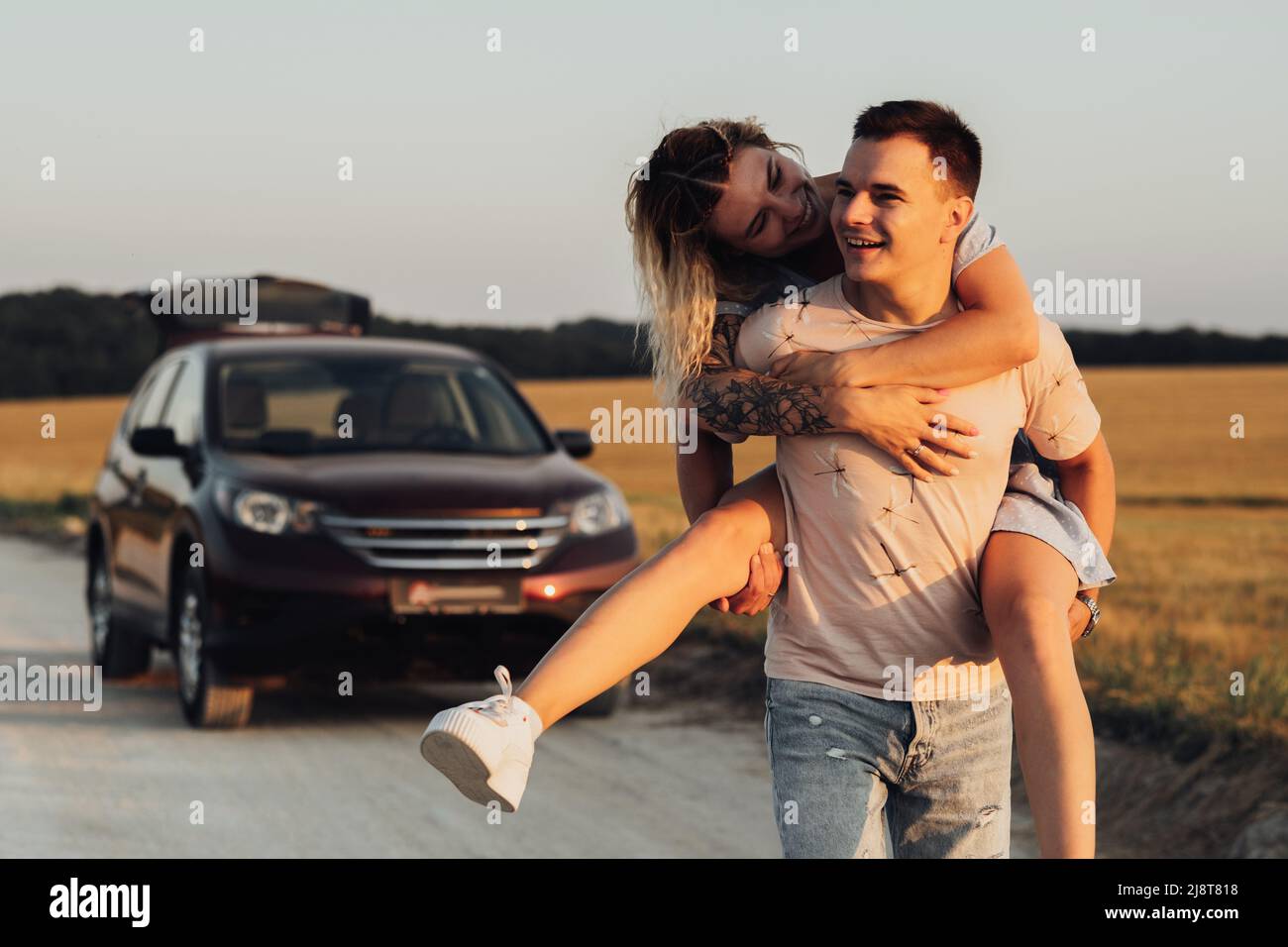 Kaukasischer Mann trägt Freundin auf seinem Rücken auf dem Hintergrund ihres SUV-Autos, Junges Paar genießt ihre Road Trip bei Sonnenuntergang Stockfoto