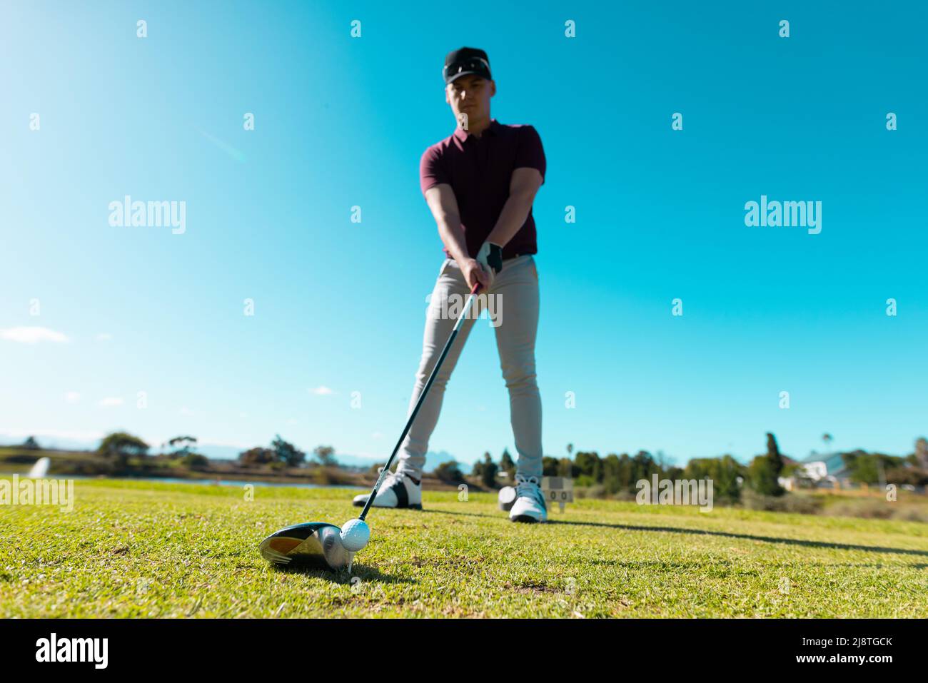 Blick aus der unteren Perspektive auf den kaukasischen jungen Mann, der den Ball mit dem Golfschläger anpeilt, während er gegen den klaren Himmel steht Stockfoto