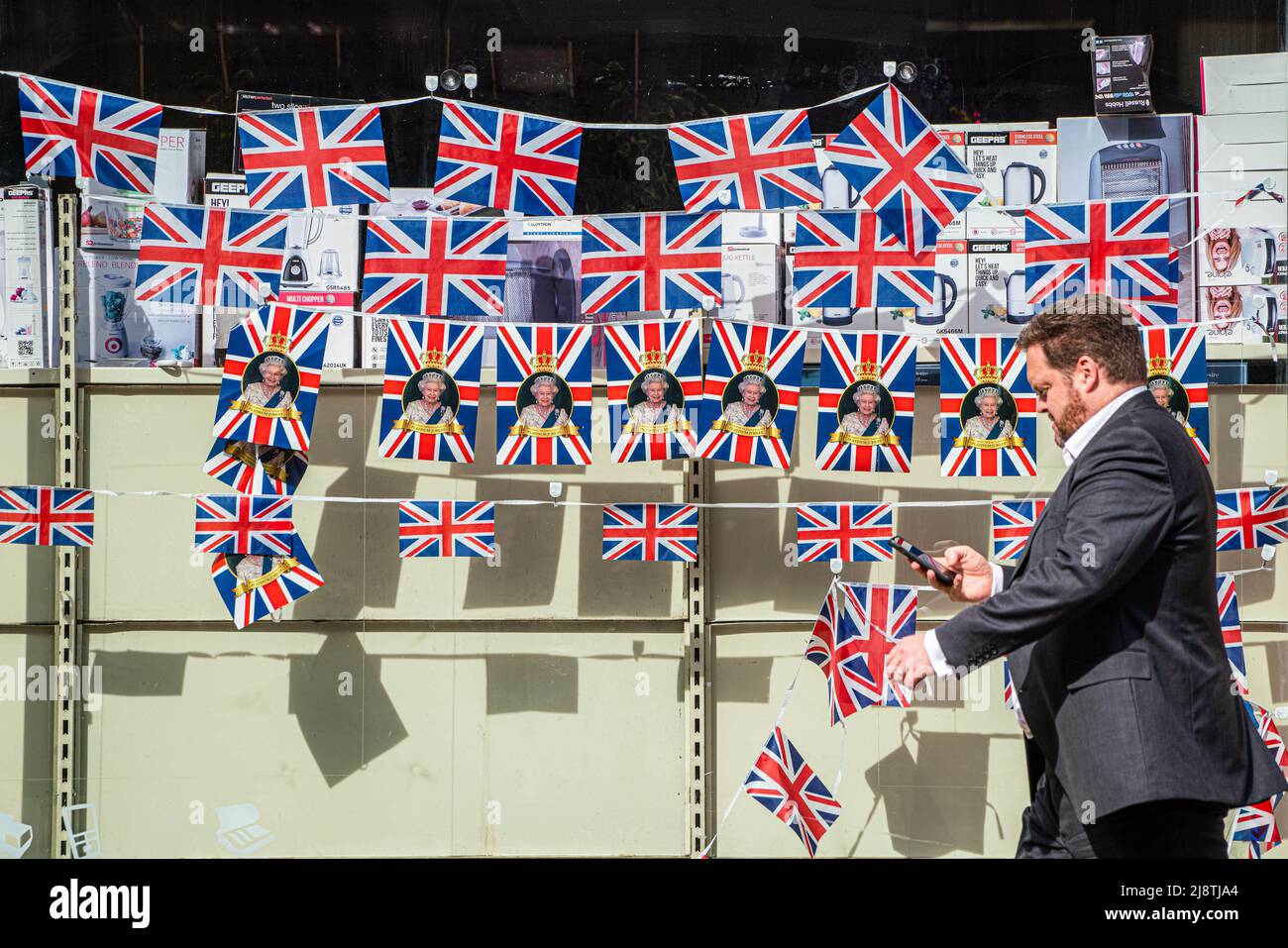 London, 18. Mai 2022. Eine Fußgängerin im Stadtzentrum von Wimbledon geht an einem Schaufenster vorbei, das mit Union Jack-Flaggen geschmückt ist und das Platinjubiläum feiert, da Königin Elizabeth 70 Jahre als die am längsten dienende britische Monarchie seit ihrer Thronbesteigung im Jahr 1952 feiert. Kredit. amer Ghazzal/Alamy Live Nachrichten Stockfoto
