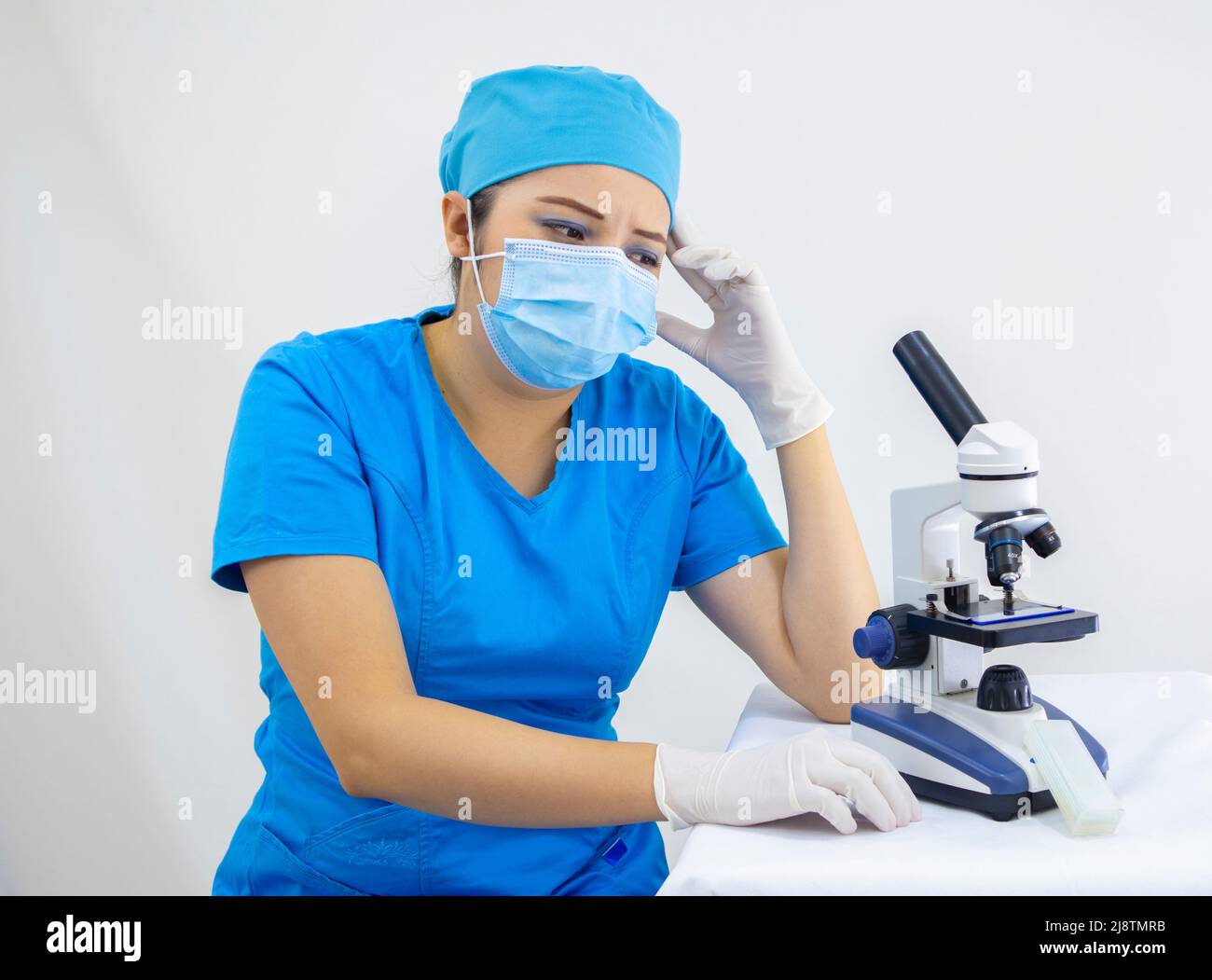 Schöne Frau Labortechniker trägt einheitliche und blaue OP-Kappe, chirurgischen Schutz, Analyse von Proben mit dem Mikroskop, auf weißem Hintergrund Stockfoto