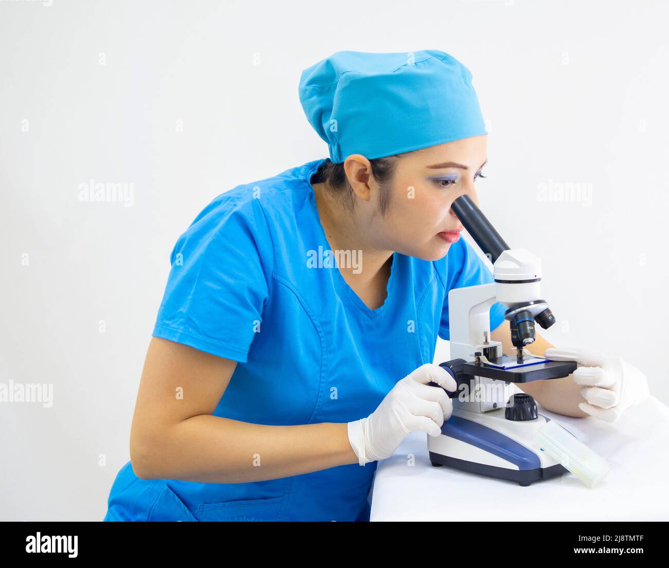Schöne Frau Labortechniker trägt einheitliche und blaue OP-Kappe, Latexhandschuhe, Analyse der Proben mit dem Mikroskop, auf weißem Hintergrund Stockfoto