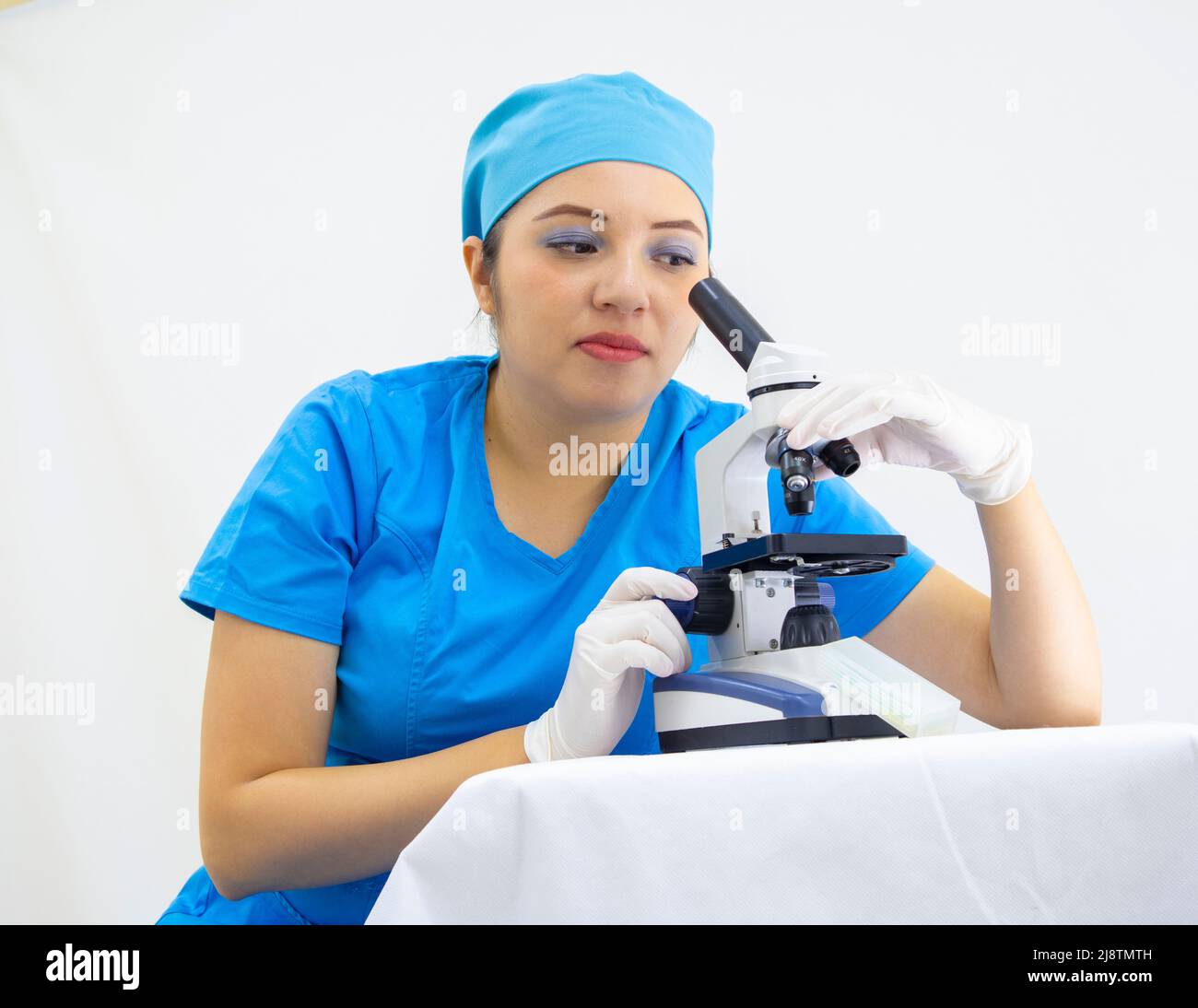 Schöne Frau Labortechniker trägt einheitliche und blaue OP-Kappe, Latexhandschuhe, Analyse der Proben mit dem Mikroskop, auf weißem Hintergrund Stockfoto