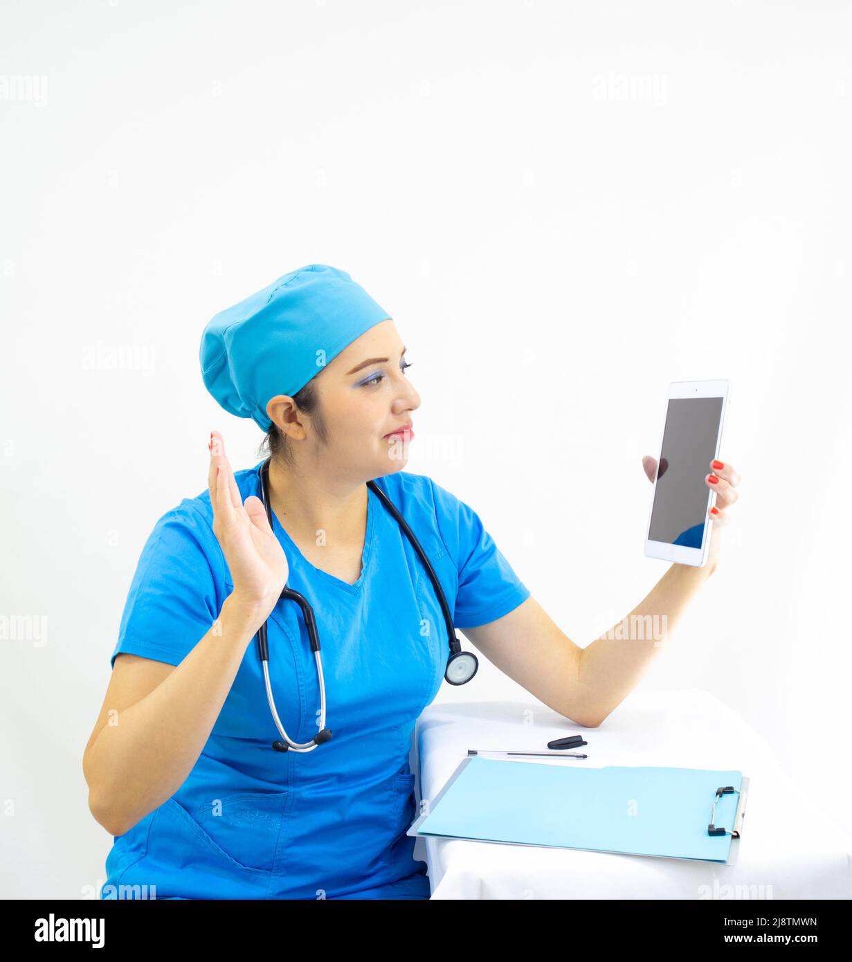 Schöne Ärztin, trägt blaue Uniform und blaue OP-Kappe, Stethoskop am Hals, winkt auf die Tablette, zeigt Ergebnisse auf weißem Hintergrund Stockfoto