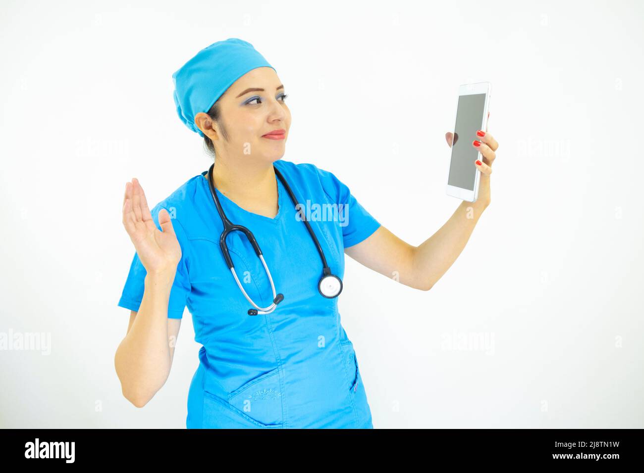 Schöne Ärztin, trägt blaue Uniform und blaue OP-Kappe, Stethoskop am Hals und winkt auf weißem Hintergrund auf die Tablette Stockfoto
