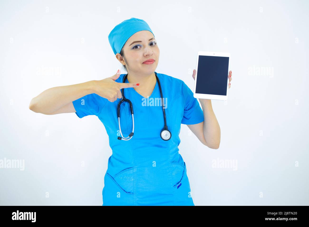 Schöne Ärztin, getragen von Uniform und blauer OP-Kappe, Stethoskop am Hals, zeigt auf die Tablette auf weißem Hintergrund Stockfoto