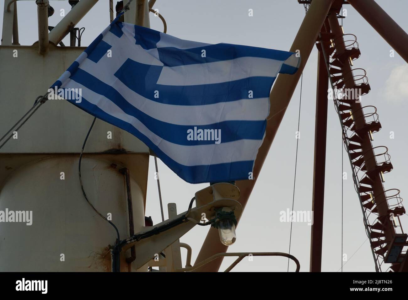 Die griechische Flagge flattert auf dem Mast des im Hafen von Piräus verankerten Handelscontainerschiffes. Stockfoto