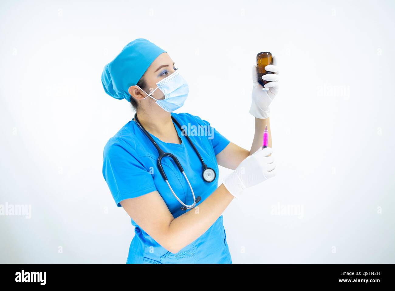 Schöne Frau Arzt trägt Maske Gesicht, einheitliche und blaue OP-Kappe, Stethoskop und Latexhandschuhe, Vorbereitung der Injektion auf weißem Hintergrund Stockfoto