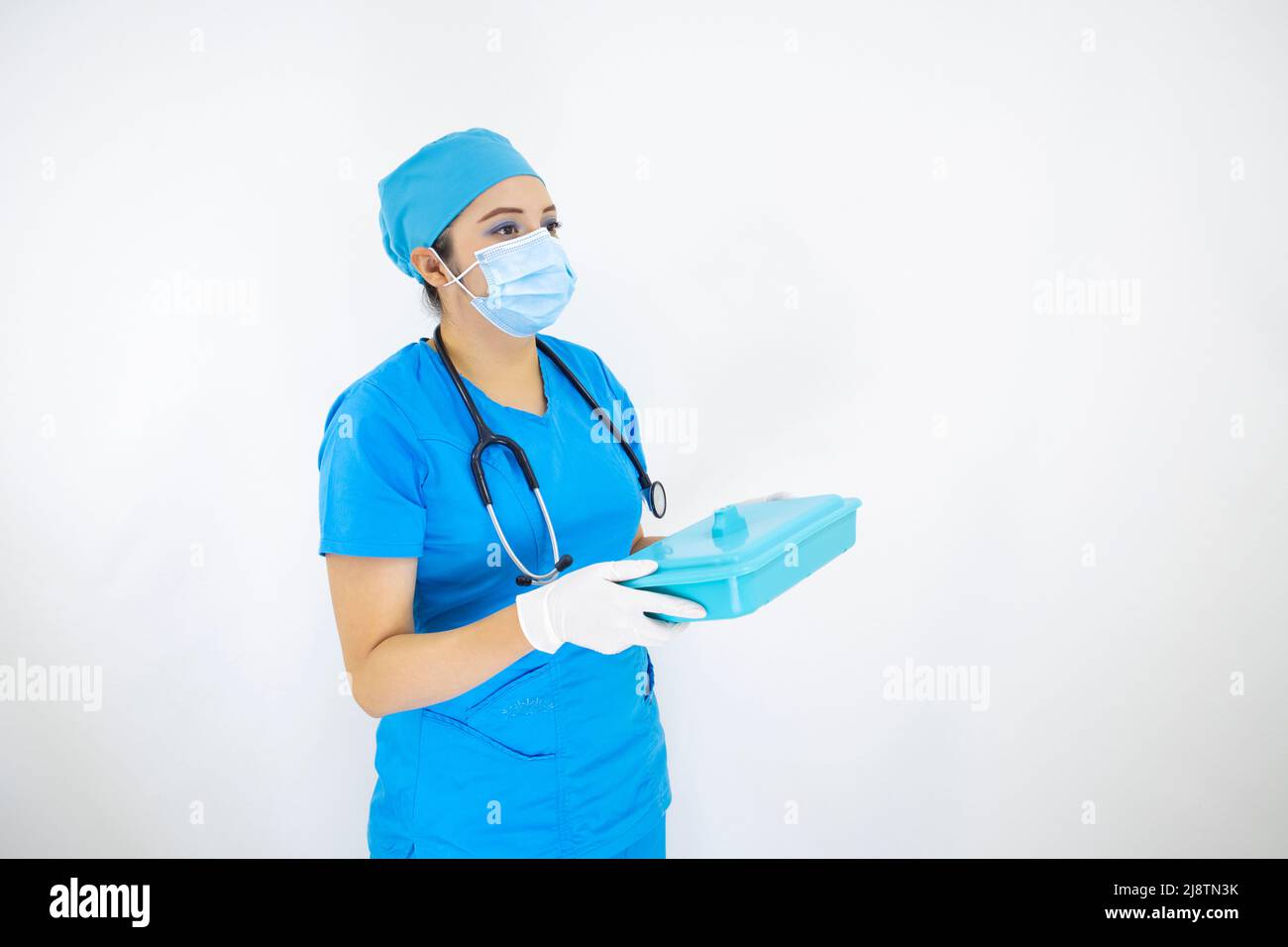 Schöne medizinische Frau trägt Gesichtsmaske, einheitliche und blaue OP-Kappe, Stethoskop und Latexhandschuhe, Vorbereitung Instrumente Fall auf weißem Hintergrund Stockfoto