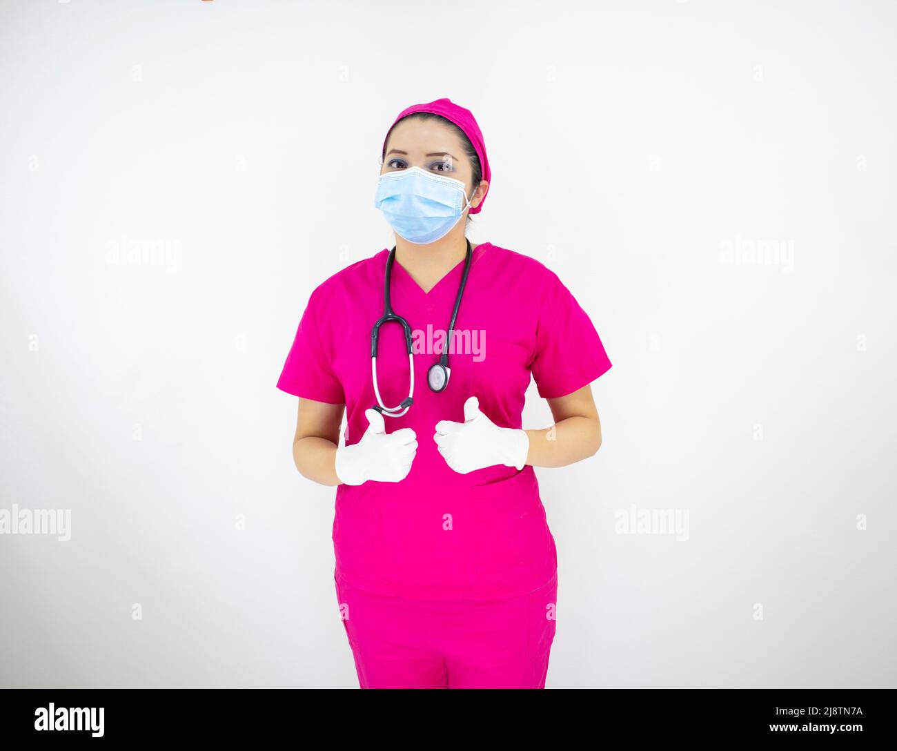 Schöne weibliche medizinische Assistentin trägt Gesichtsmaske, rosa Uniform und rosa OP-Kappe, Latexhandschuhe und Stethoskop, auf weißem Hintergrund Stockfoto