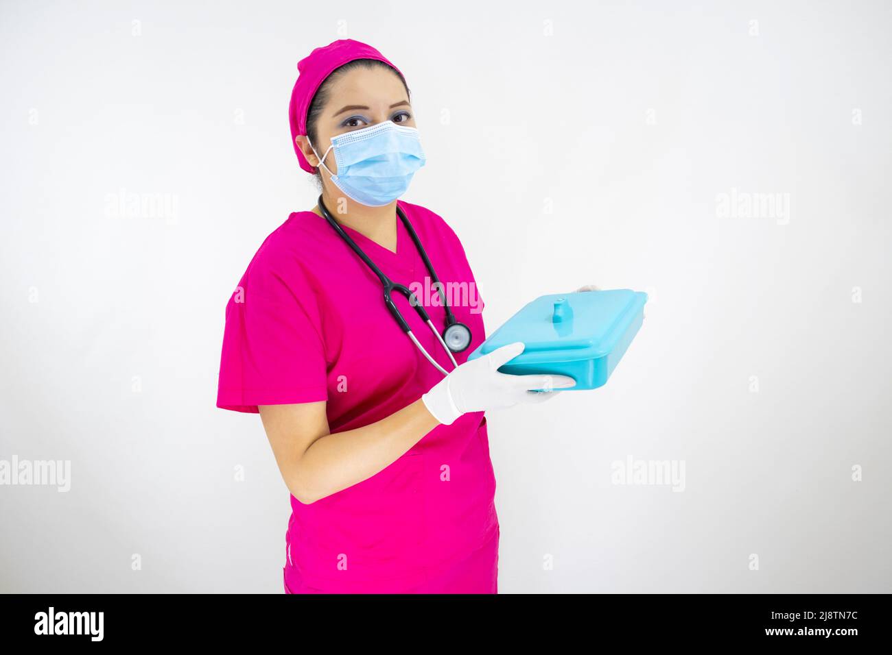 Schöne weibliche medizinische Assistentin trägt Gesichtsmaske, rosa Uniform und rosa OP-Kappe, Stethoskop, Vorbereitung Instrumente Fall auf weißem Hintergrund Stockfoto