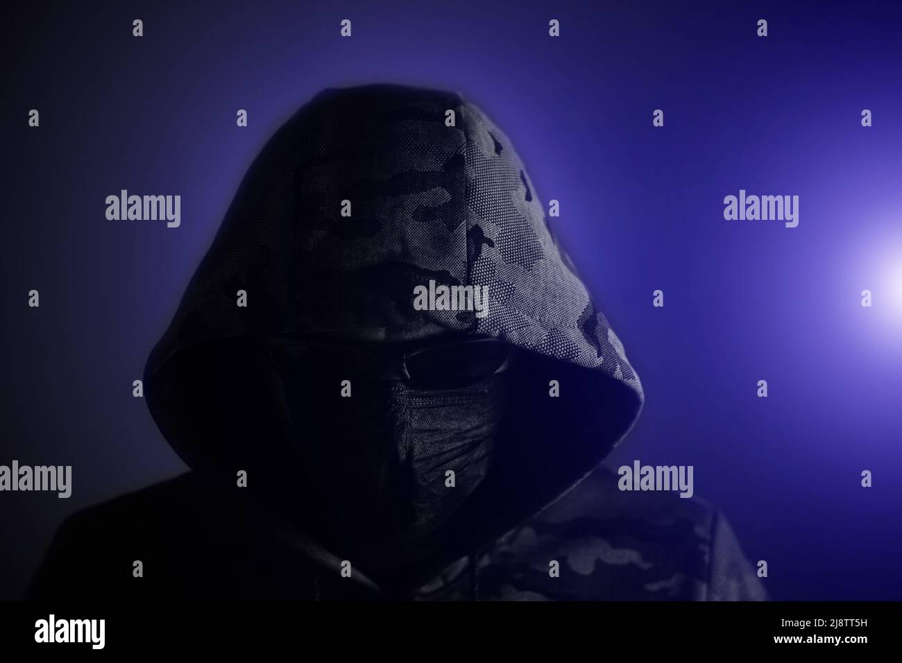 Stilvoller Mann in dunklem Hoodie, schwarzer Brille und dunkelem Hintergrund in blauem Licht, Hacker, Angreifer und Bandit Stockfoto
