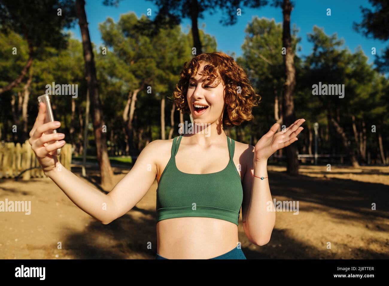 Glückliche Rotschopf-Frau, die online in einer Videokonferenz spricht oder Selfie-Videos für soziale Medien mit einem Smartphone aufzeichnet, während sie auf dem Stadtpark steht, ou Stockfoto