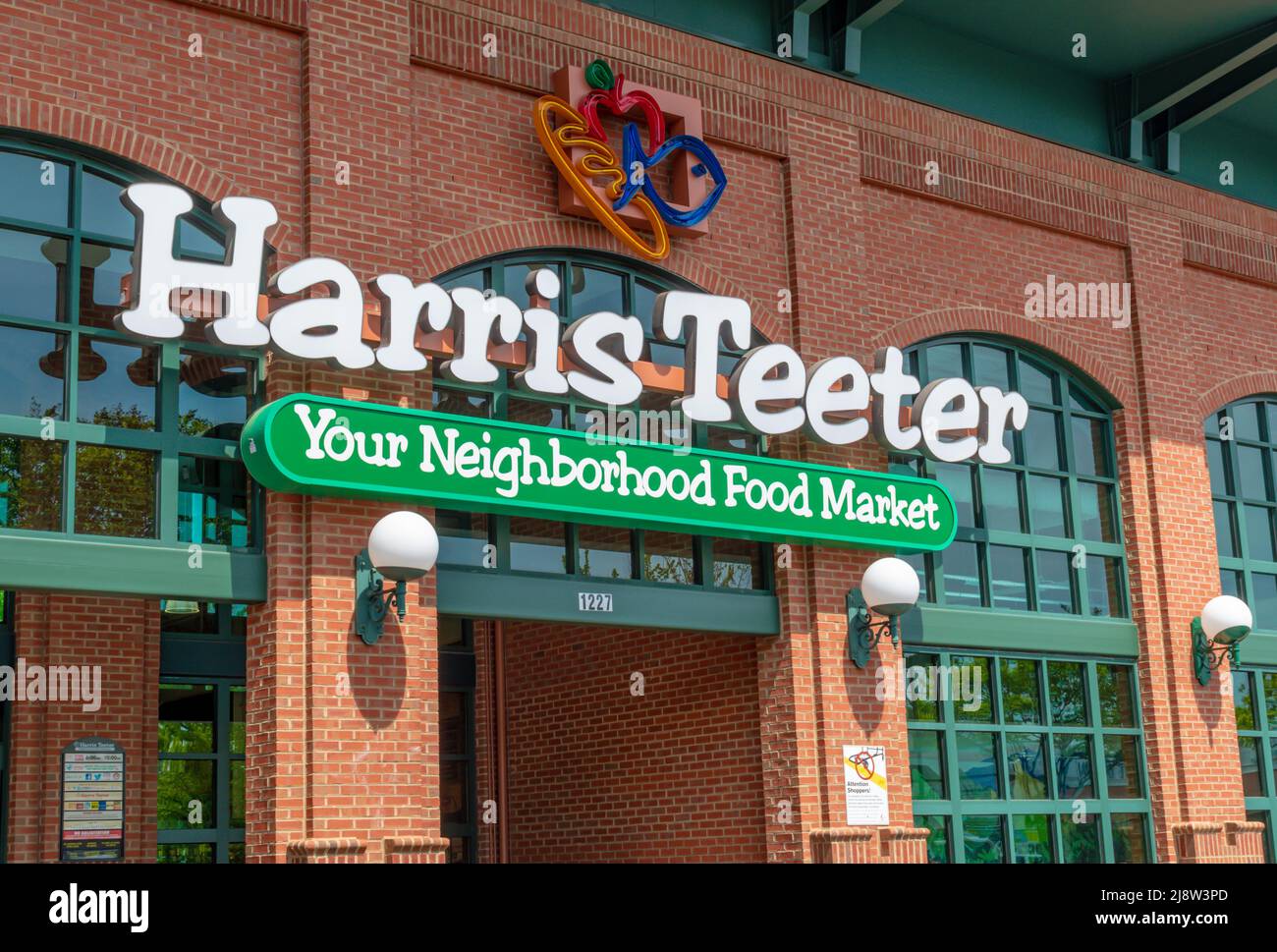 „Harris Teeter“-Außenfassade mit Marken- und Logo-Beschilderung mit farbenfrohem Neon-Logo, reflektierenden Glasfenstern, Globenleuchtern neben dem Eingang im Sonnenlicht. Stockfoto