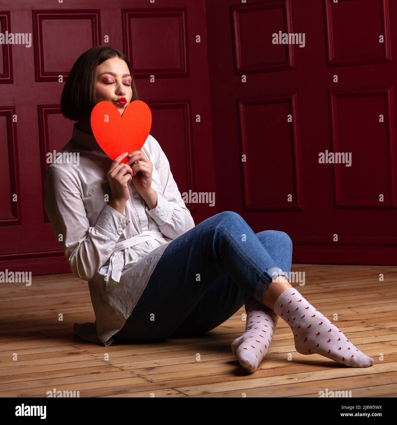 Kurzhaarige Frau mit geschlossenen Augen und Lippen Formen Kuss sitzen auf Holzboden Beine gekreuzt und roten Hintergrund halten rote Herzform vor Stockfoto