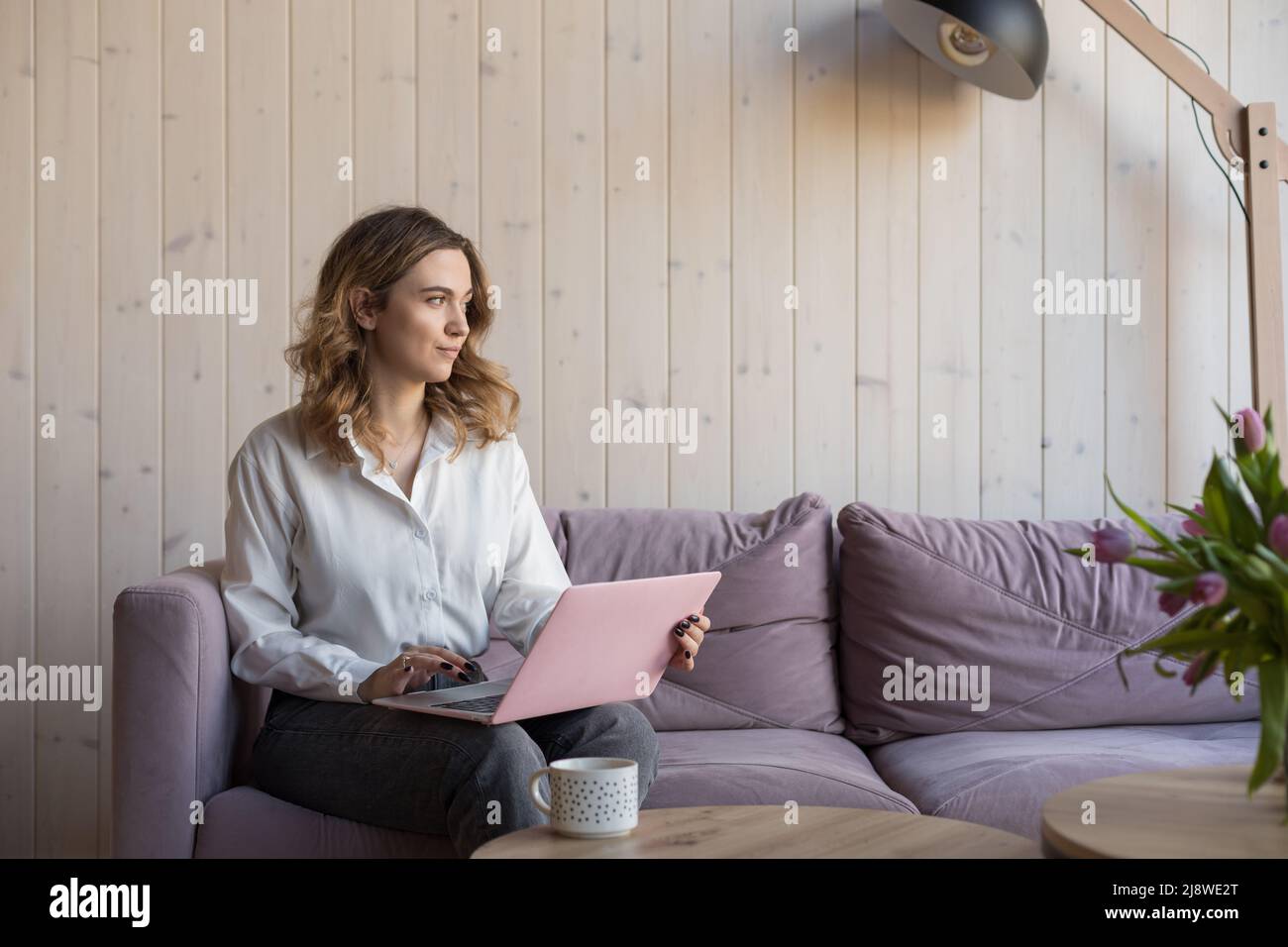 Junge seriöse braunhaarige Frau arbeitet zu Hause online auf der Couch sitzend, reflektiert sie auf einem Brief an den Kunden, aus dem Fenster blickend. Online zu Hause arbeiten Stockfoto