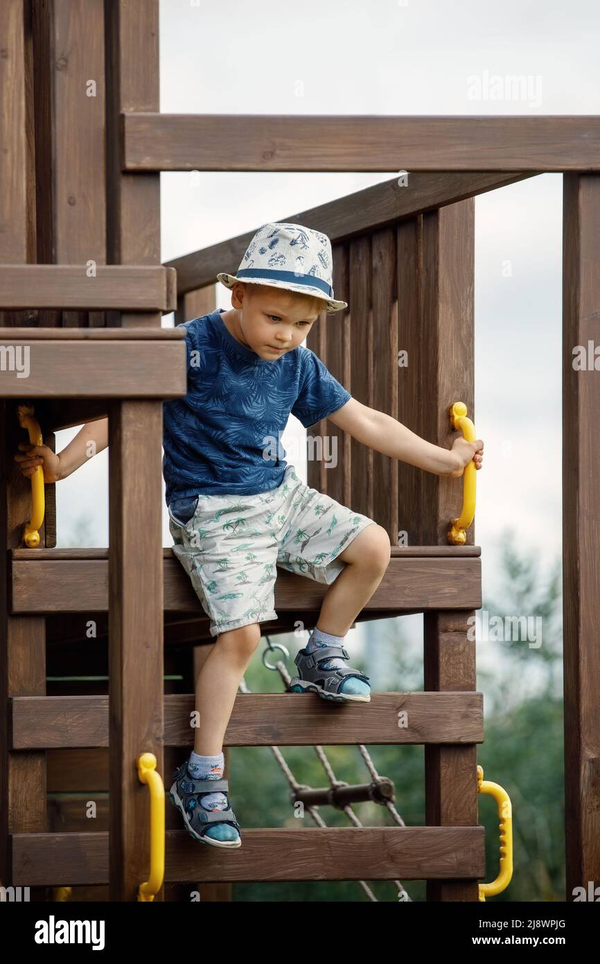 Der Junge spielt auf einem Spielplatz im Freien, er klettert gefährlich auf eine Holzleiter. Ein aktives Kind braucht elterliche Fürsorge für die Sicherheit. Stockfoto