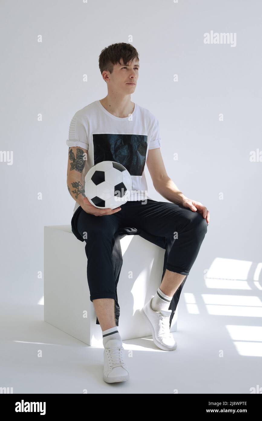 Stilvoller Kaukasier mit tätowiertem Arm, der den Fußball hält und in die Ferne blickt, während er im weißen Raum auf einem Würfel sitzt Stockfoto