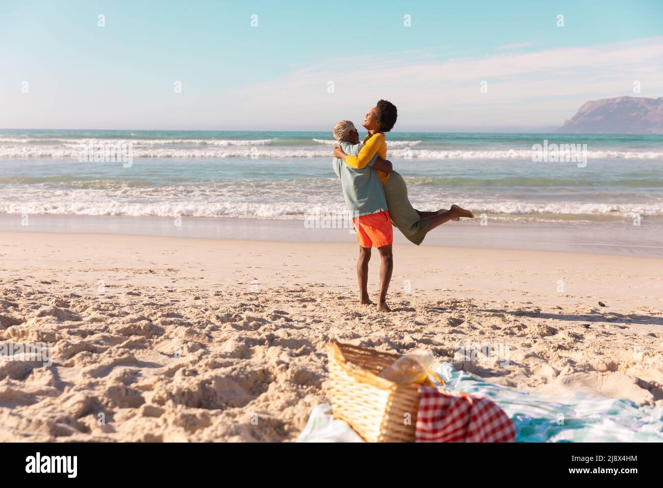 Glücklicher afroamerikanischer älterer Mann, der eine reife Frau trägt, während er am Strand gegen Meer und Himmel steht Stockfoto