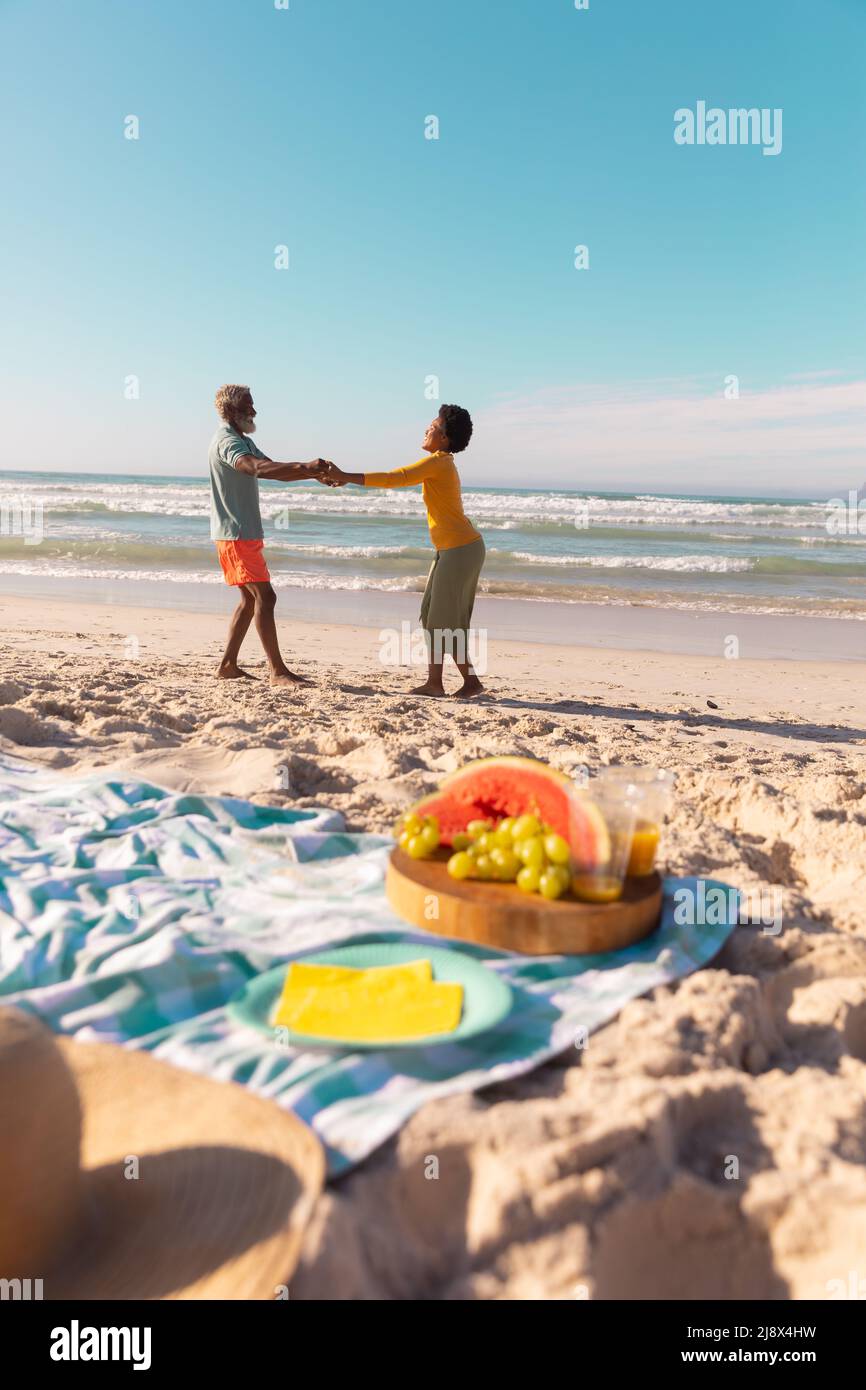 Seitenansicht eines afroamerikanischen Paares, das die Hände hält und am Sandstrand vor blauem Himmel tanzt Stockfoto