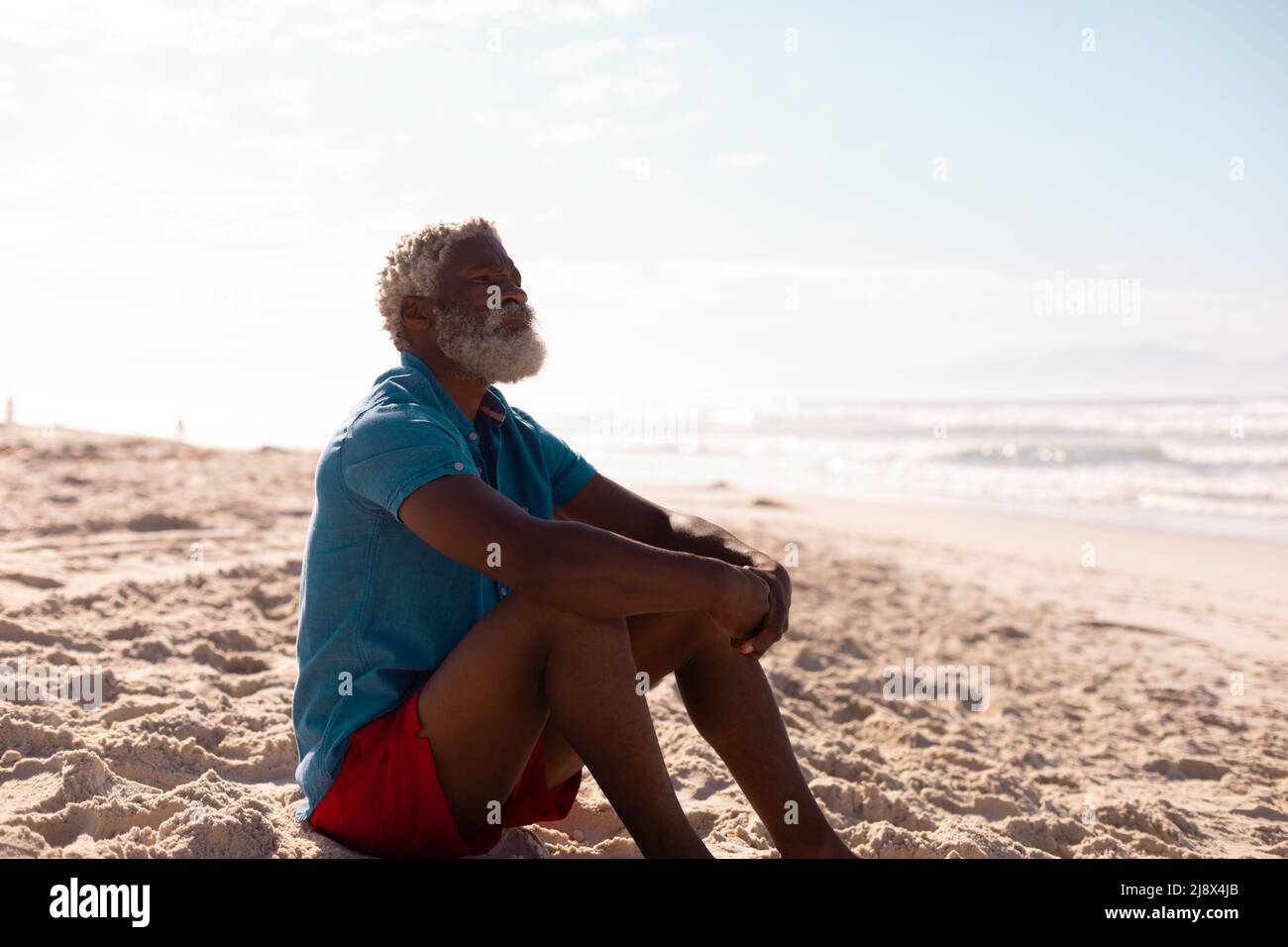 Nachdenklicher bärtiger afroamerikanischer Senioren mit grauen Haaren, der am Sandstrand gegen den Himmel sitzt Stockfoto