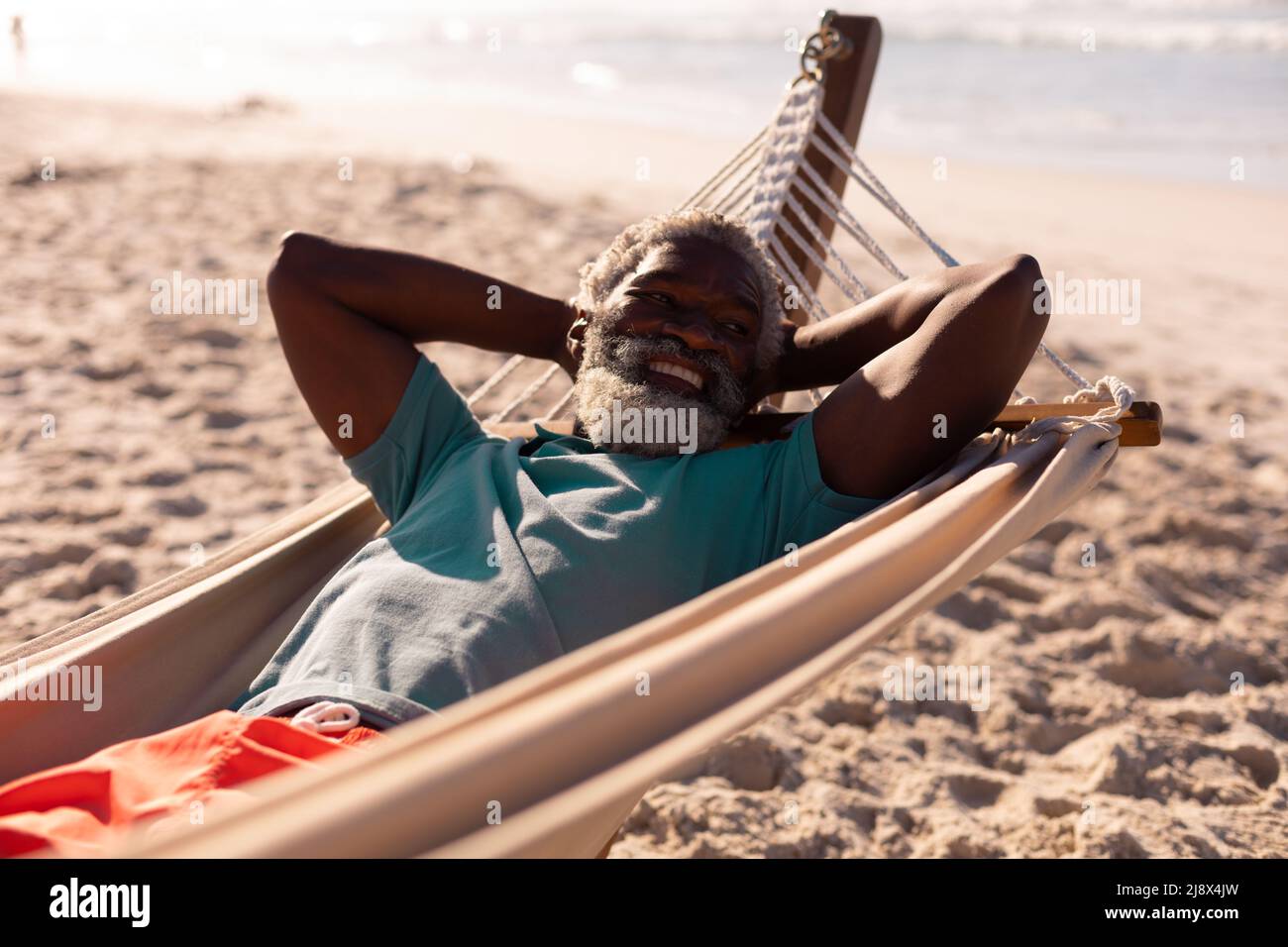 Bärtiger afroamerikanischer Senior mit Händen hinter dem Kopf, der im Sommer am Strand in einer Hängematte liegt Stockfoto