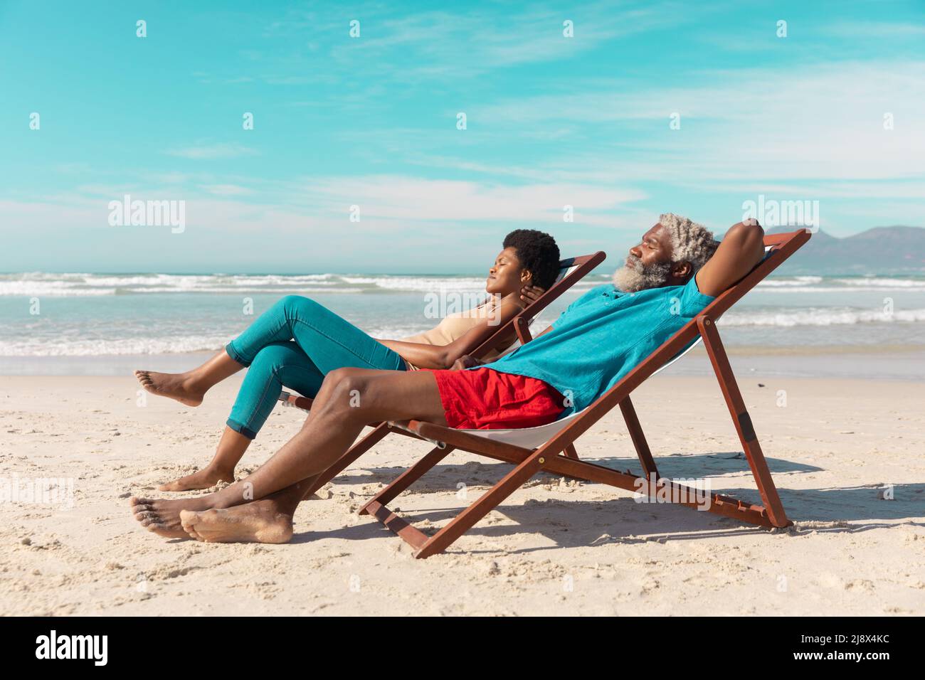 Seitenansicht eines afroamerikanischen Paares, das sich im Sommer auf Liegestühlen vor dem Meer und dem blauen Himmel entspannen kann Stockfoto