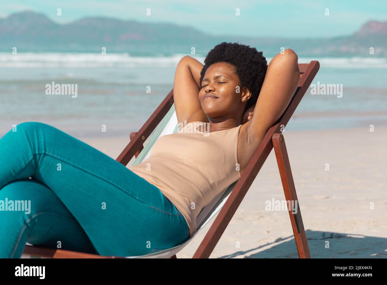 afroamerikanische reife Frau mit den Händen hinter dem Kopf und geschlossenen Augen beim Entspannen auf dem Liegestuhl am Strand Stockfoto