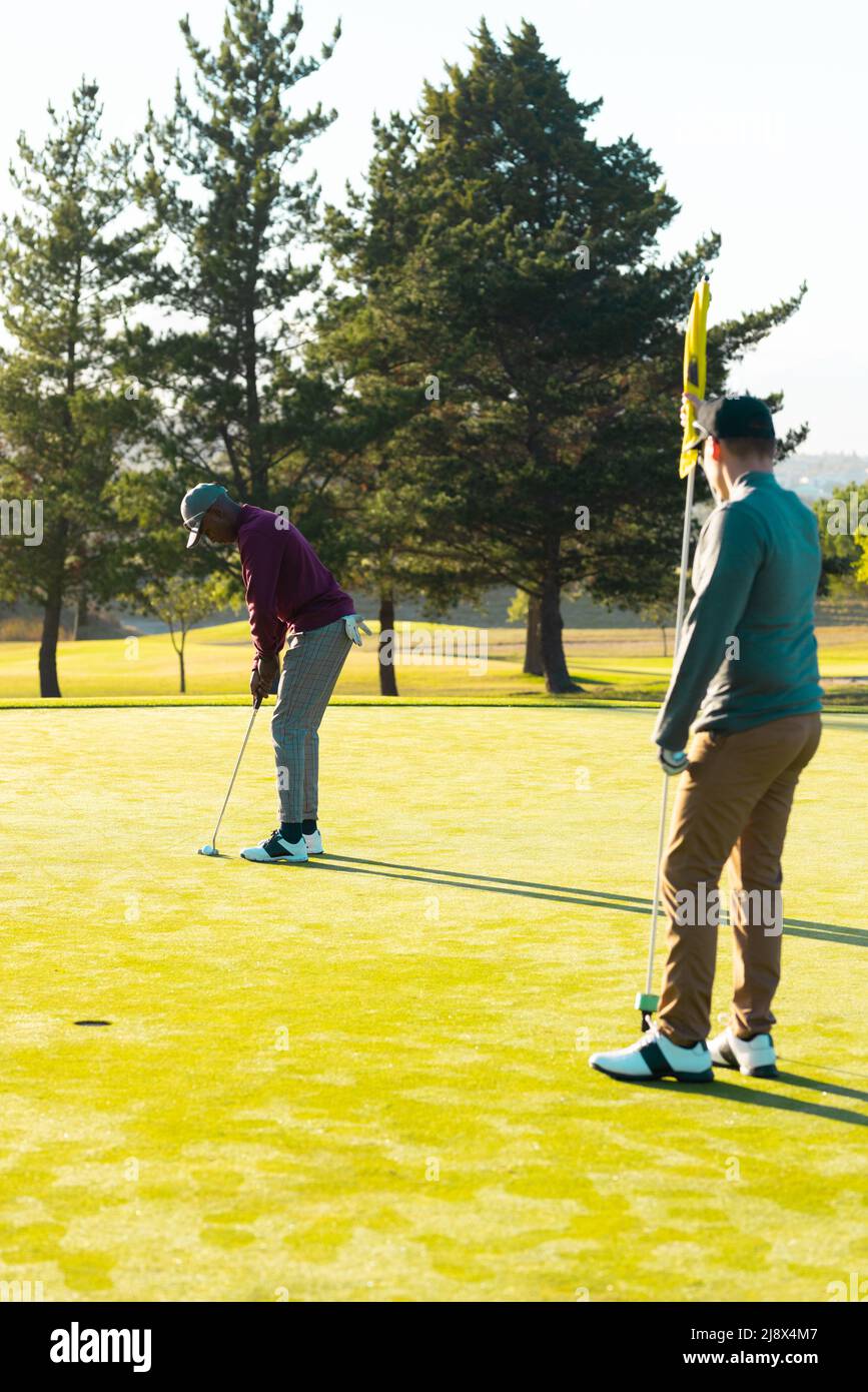 Kaukasischer junger Mann mit Golfflagge und afroamerikanischem Freund, der Golfball mit Schläger auf dem Golfplatz schlägt Stockfoto