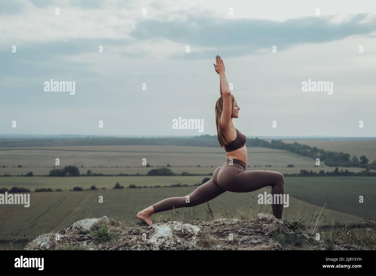 Sportliche Mädchen machen Dehnübungen auf einem Hügel, junge Frau üben Yoga im Freien mit Panoramablick nach Sonnenuntergang Stockfoto