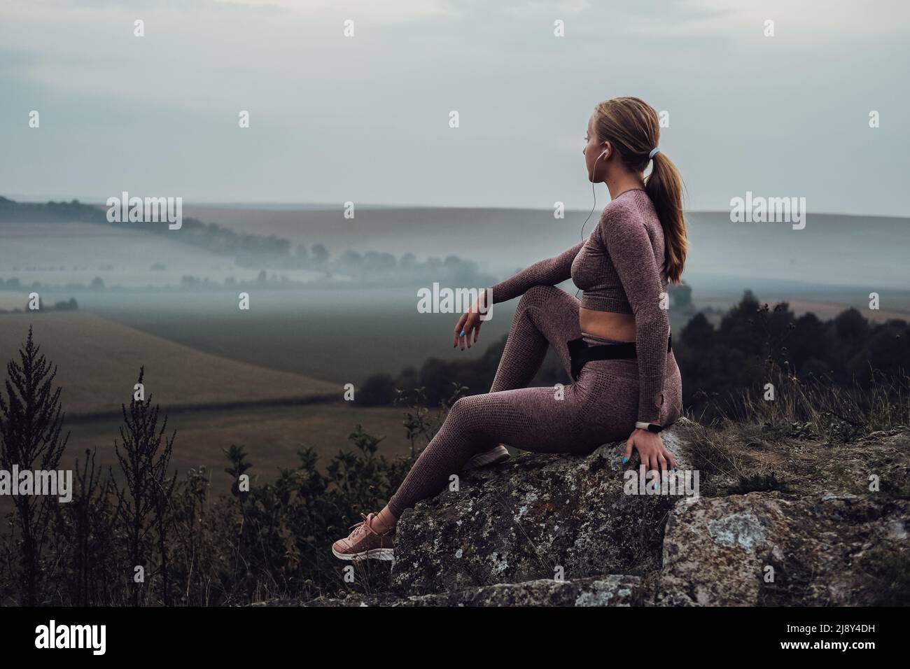Sportliche junge Frau, die auf dem Felsen auf dem Hügel sitzt, Musik in den Kopfhörern hören und nach Sonnenuntergang auf den Horizont blicken Stockfoto