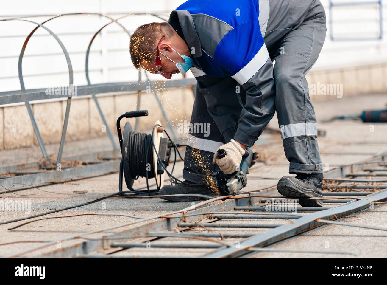 Builder in Overalls lehnt sich über und schneidet Metallblech mit Winkelschleifer und Funken fliegen. Arbeiter mit Brille bei der Arbeit auf der Straße. Authentischer Workflow. Stockfoto
